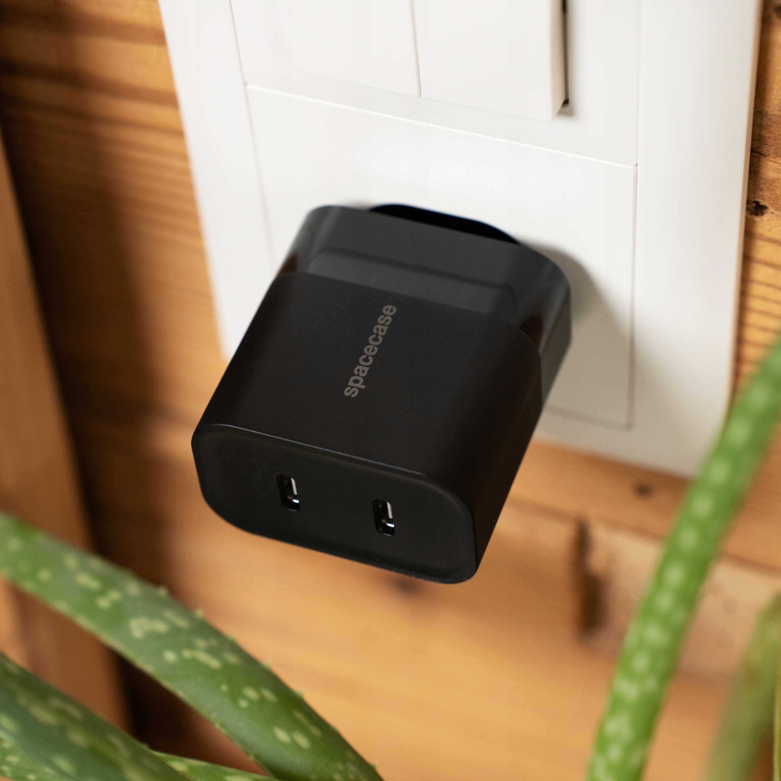 Czarna ładowarka Spacecase jest podłączona do białego gniazdka na drewnianej ścianie, z aloesem na pierwszym planie.