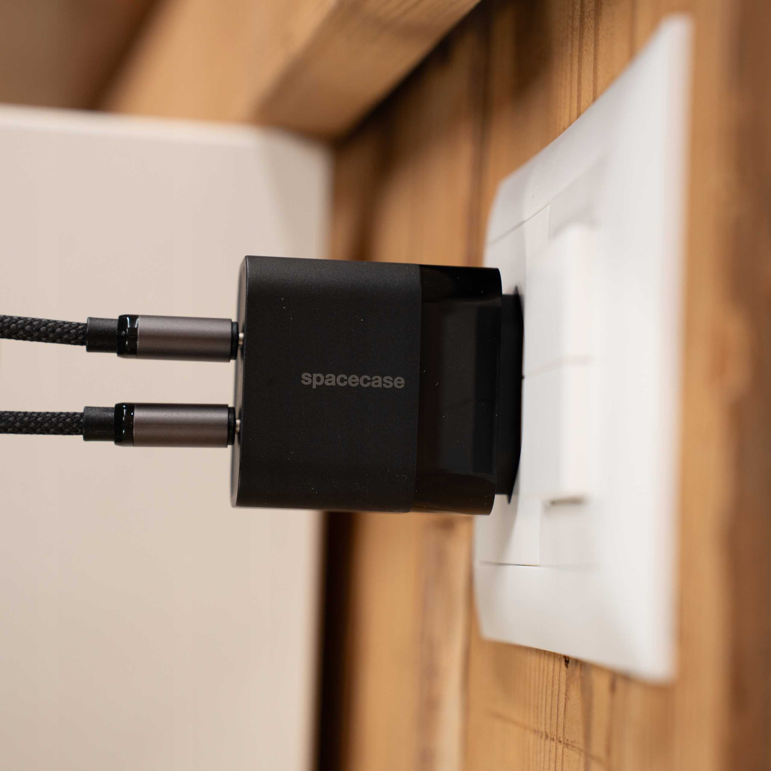 Czarna ładowarka wpięta w białe gniazdko. Dwa kable z szarymi końcami wychodzą z ładowarki. Drewniana ściana.