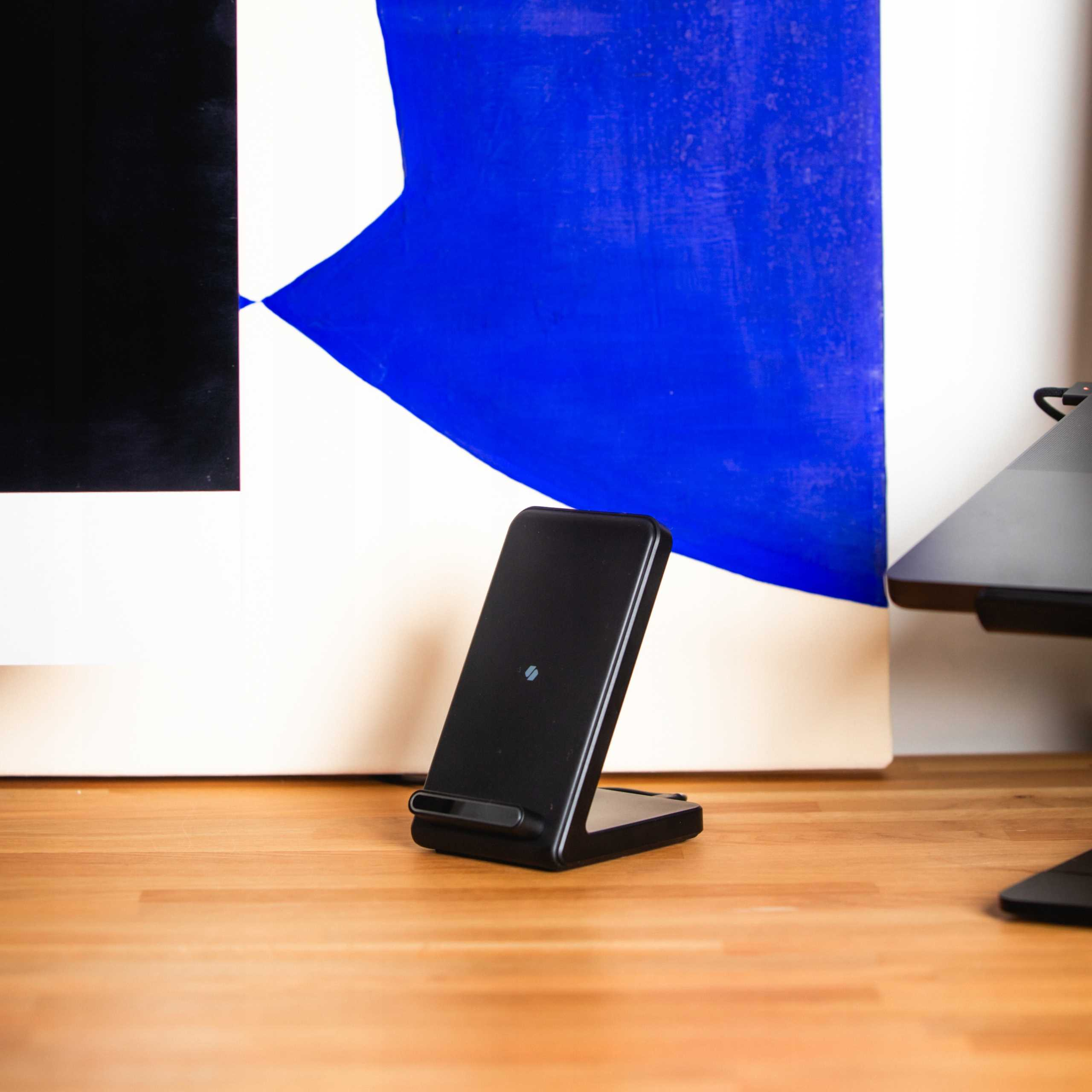 Czarna ładowarka bezprzewodowa stoi na drewnianym biurku. W tle abstrakcyjny obraz w kolorze niebiesko-białym.