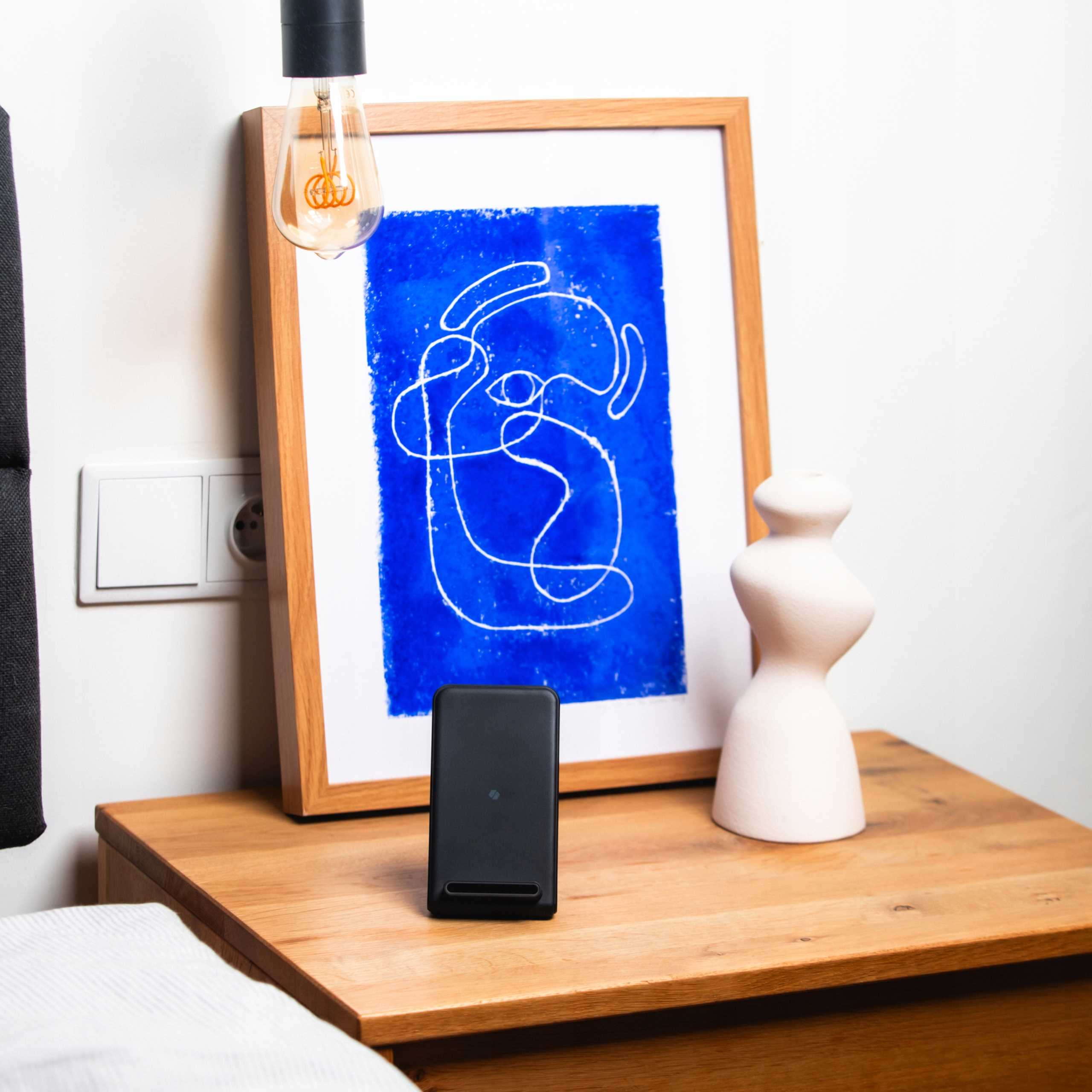 Czarne urządzenie na drewnianej szafce nocnej, oprawione niebieskie abstrakcyjne dzieło sztuki i biały przedmiot dekoracyjny. Lampa wisząca.