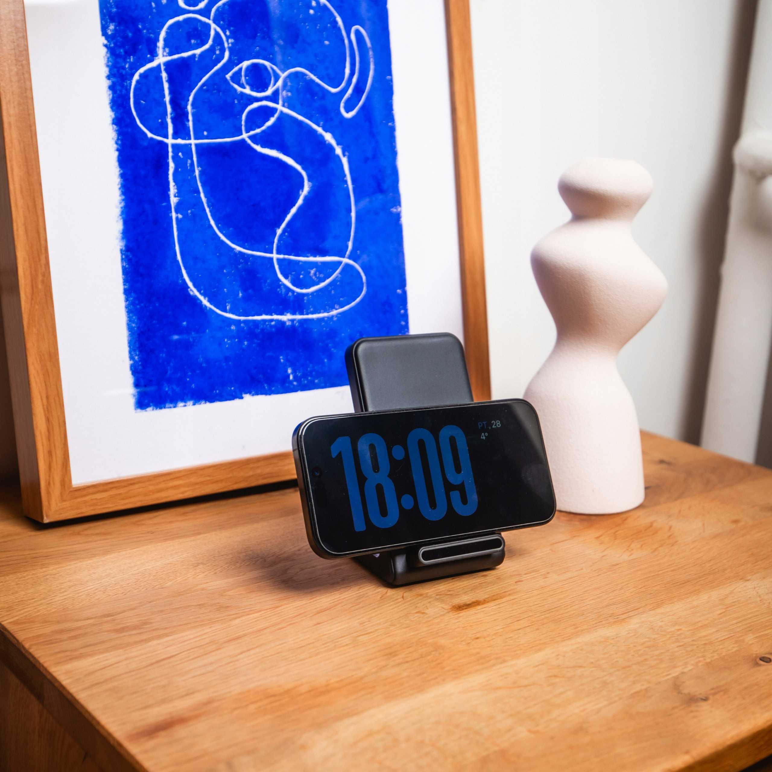 Szafka nocna z telefonem pokazującym 18:09, grafiką i wazonem. Grafika ma niebieskie tło i białe linie.