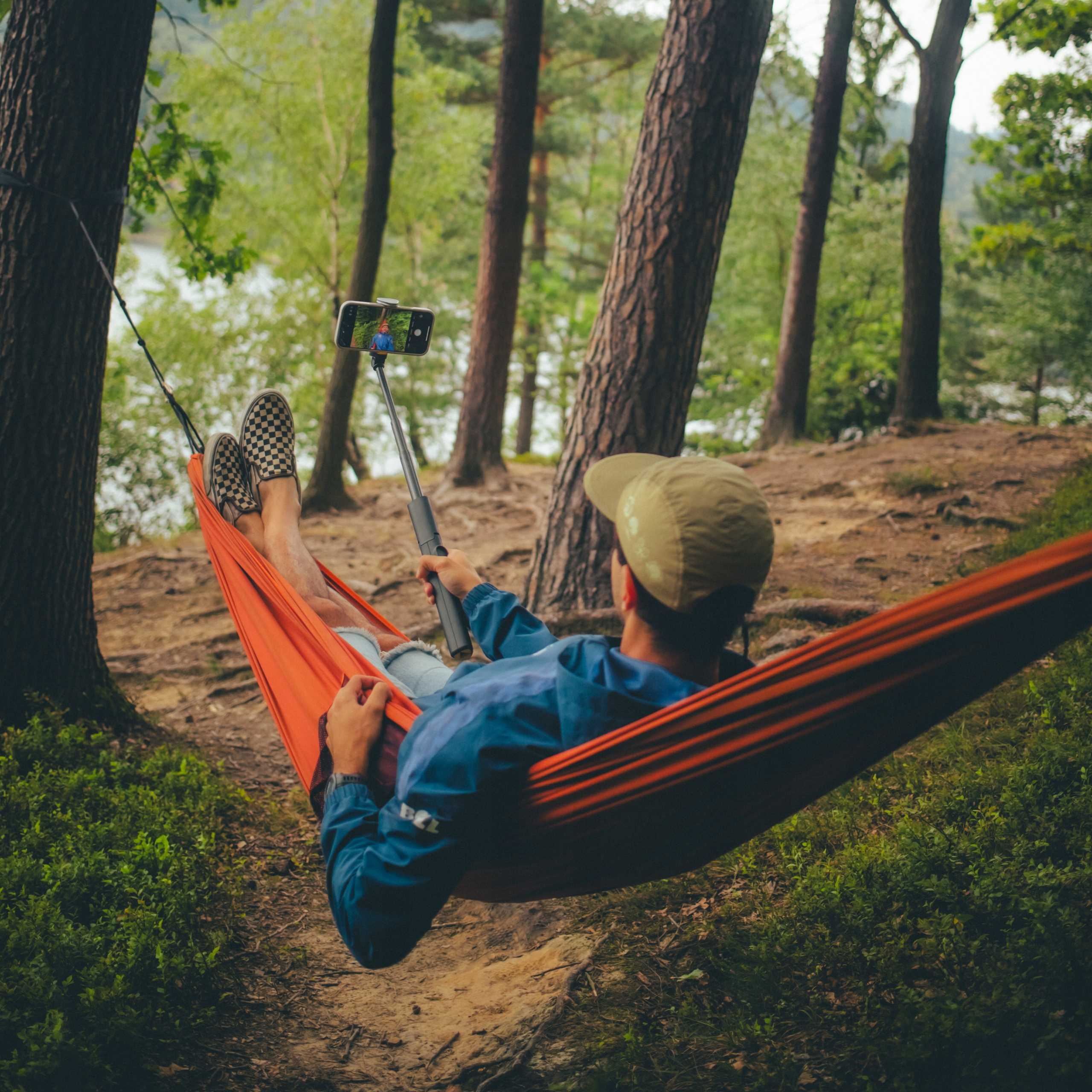 Osoba w pomarańczowym hamaku robiąca selfie telefonem. Scena w lesie z drzewami i zielonymi liśćmi.