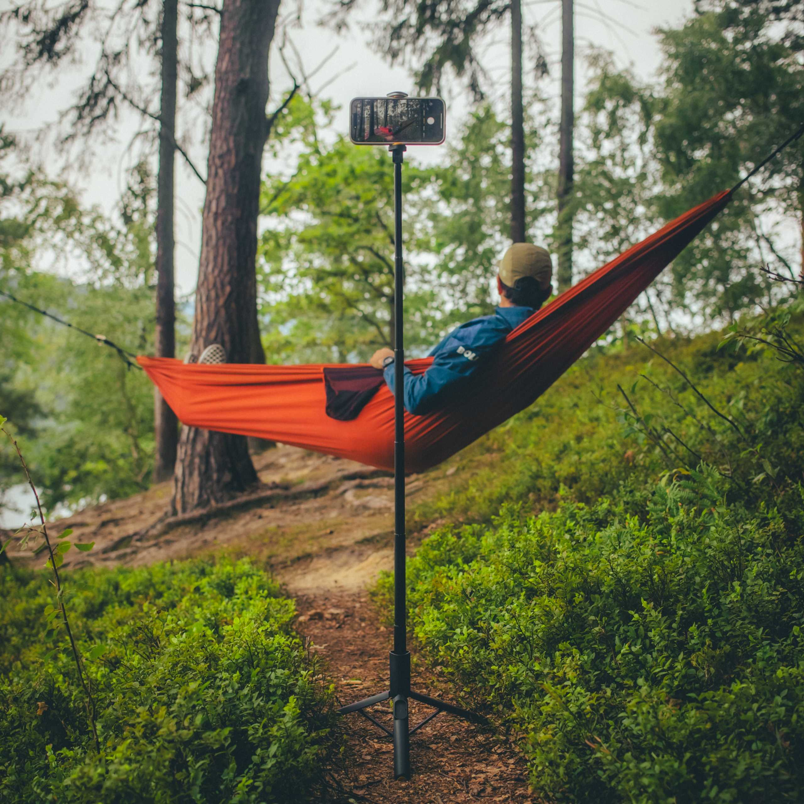 Osoba w pomarańczowym hamaku, filmująca telefonem na statywie w lesie. Otaczają krzewy i drzewa.