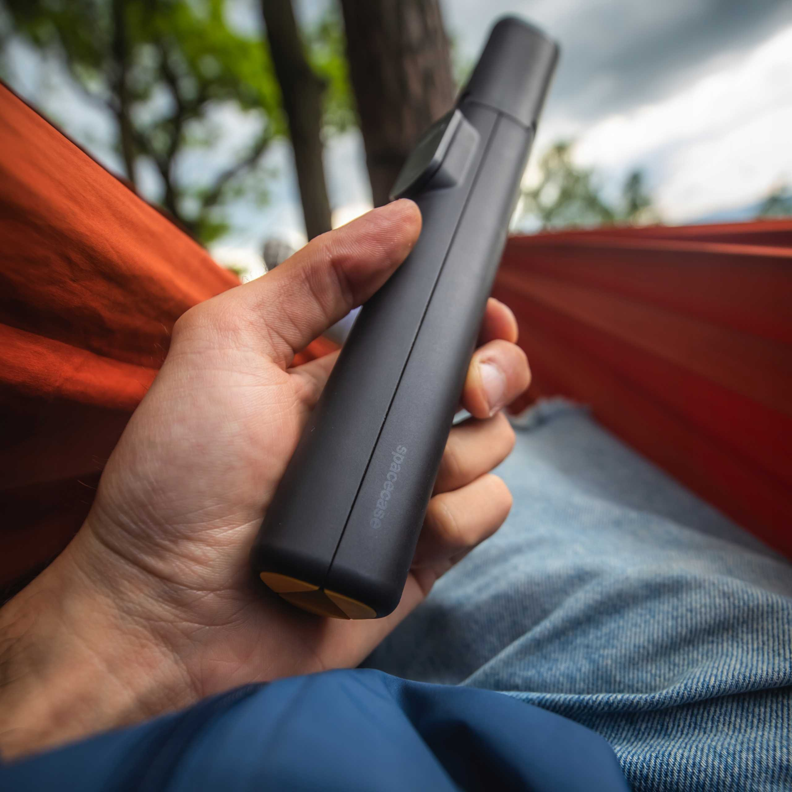 Ręka trzymająca czarne urządzenie Spacecase. Pomarańczowy hamak w tle, z drzewami i błękitnym niebem.