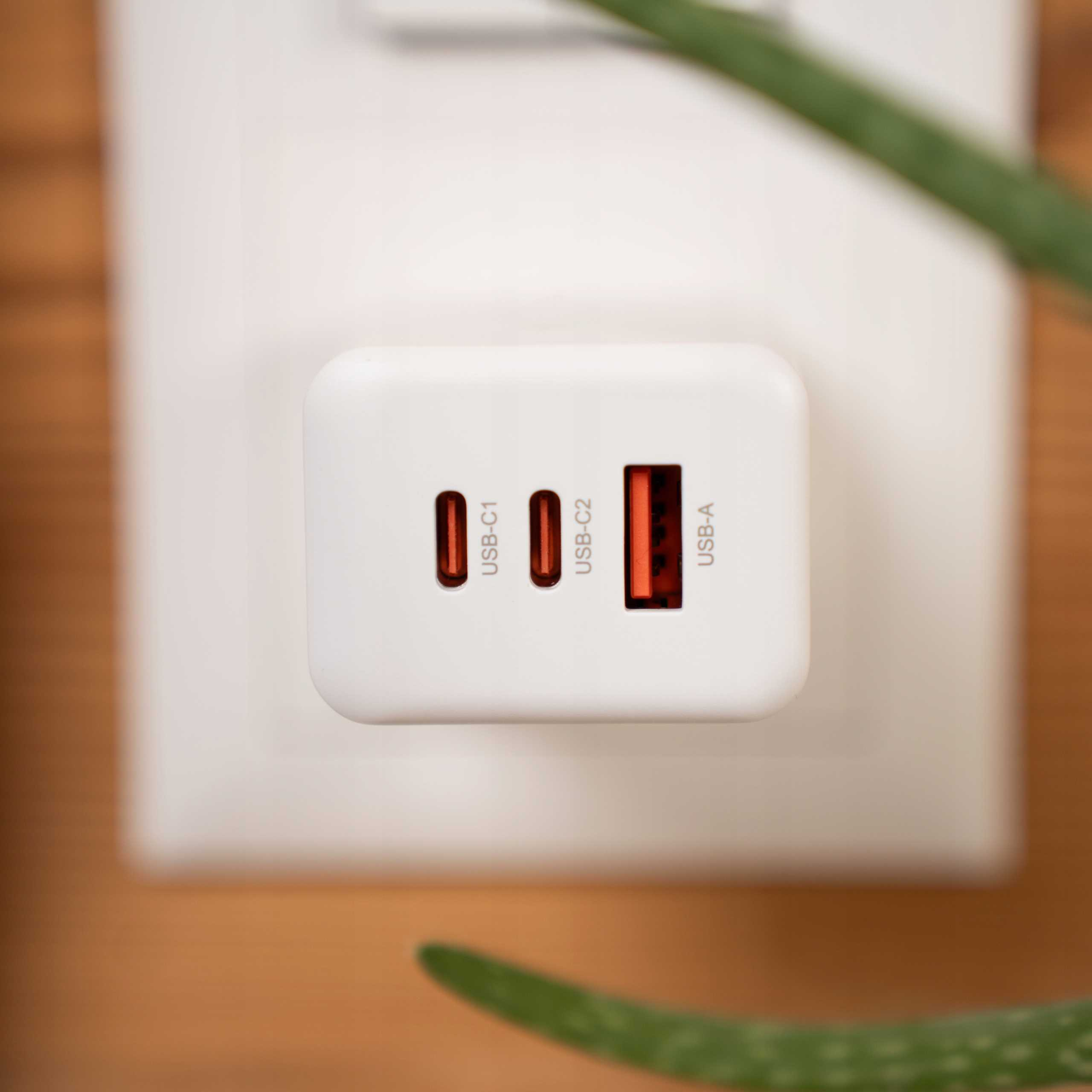 Biała ładowarka USB z trzema portami, na białej ścianie. Zielona roślina w rogu.