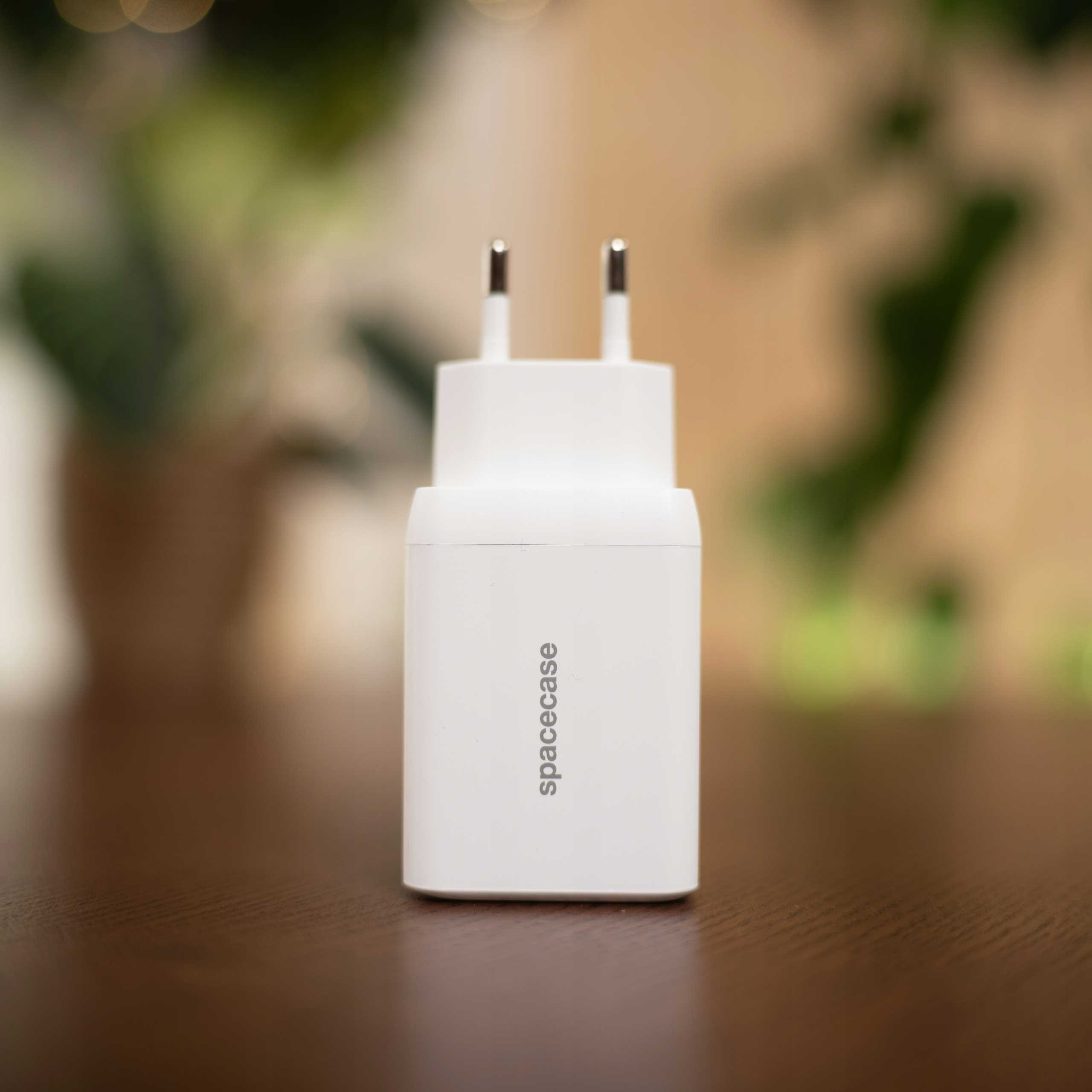 Biała ładowarka Spacecase z dwoma bolcami stoi na brązowym stole. Zielone liście są rozmyte w tle.