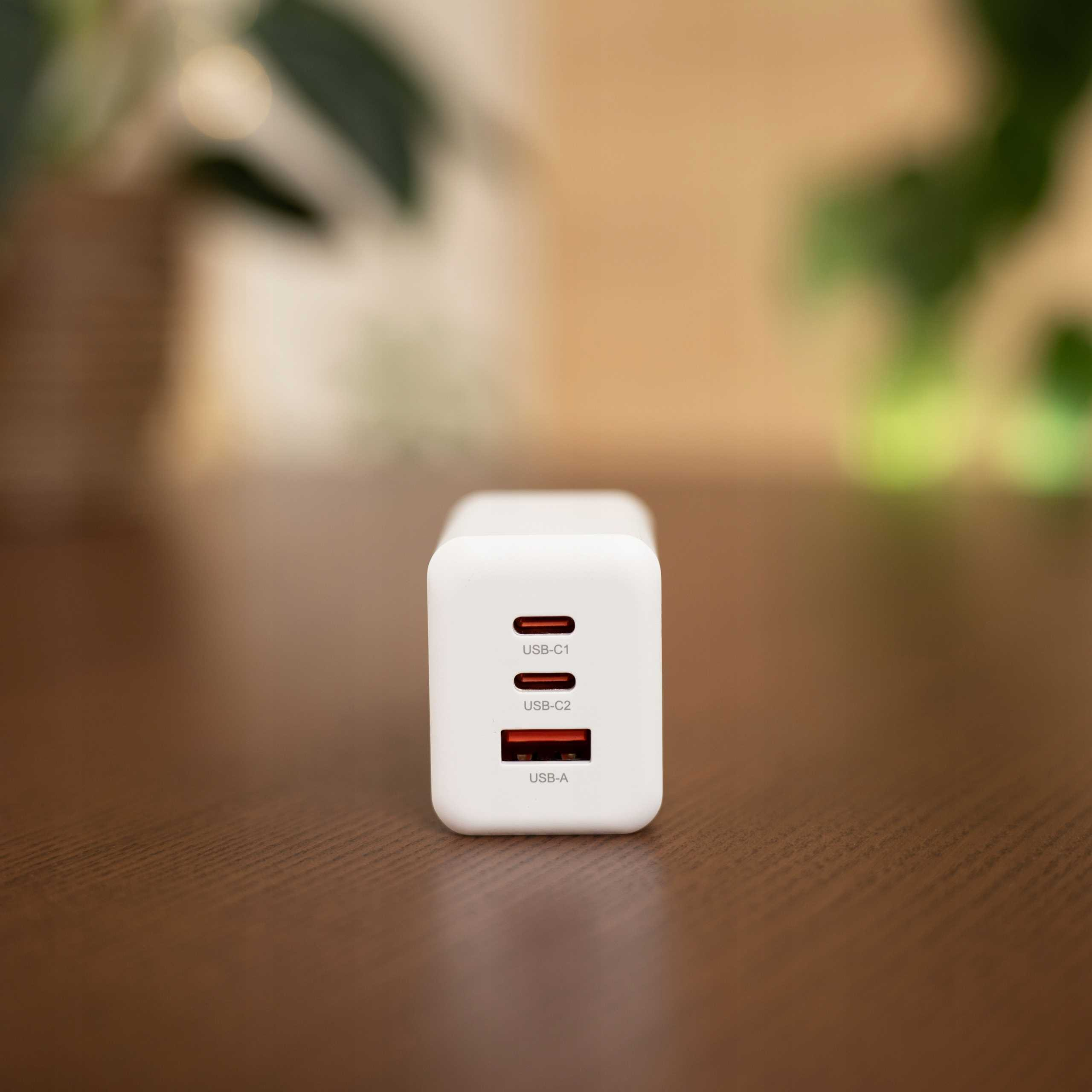 Biała ładowarka USB z trzema portami: USB-C1, USB-C2 i USB-A. Leży na drewnianym stole z rozmytym zielonym tłem.