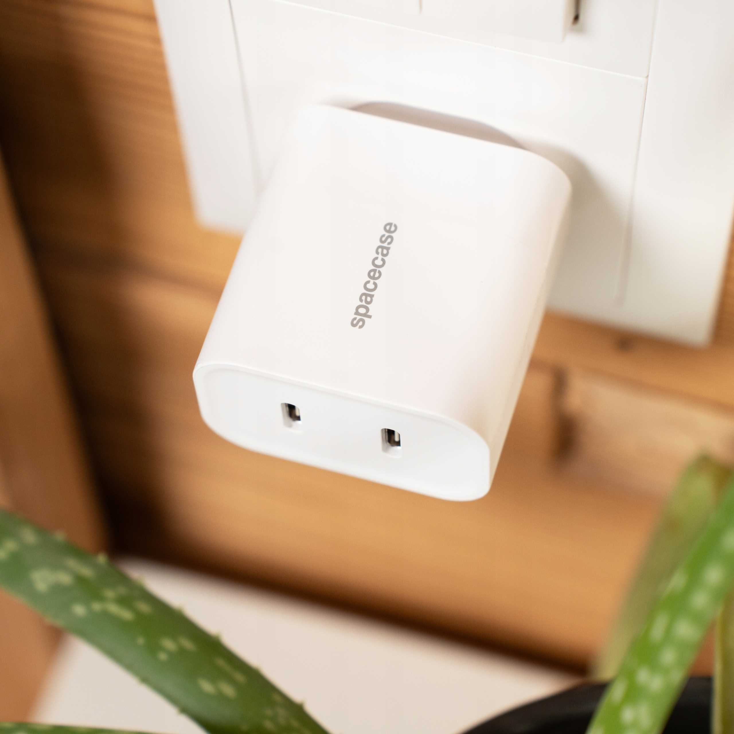 Biała ładowarka 'spacecase' jest podłączona do białego gniazdka. Brązowe drewno za gniazdkiem i zielona roślina na dole.