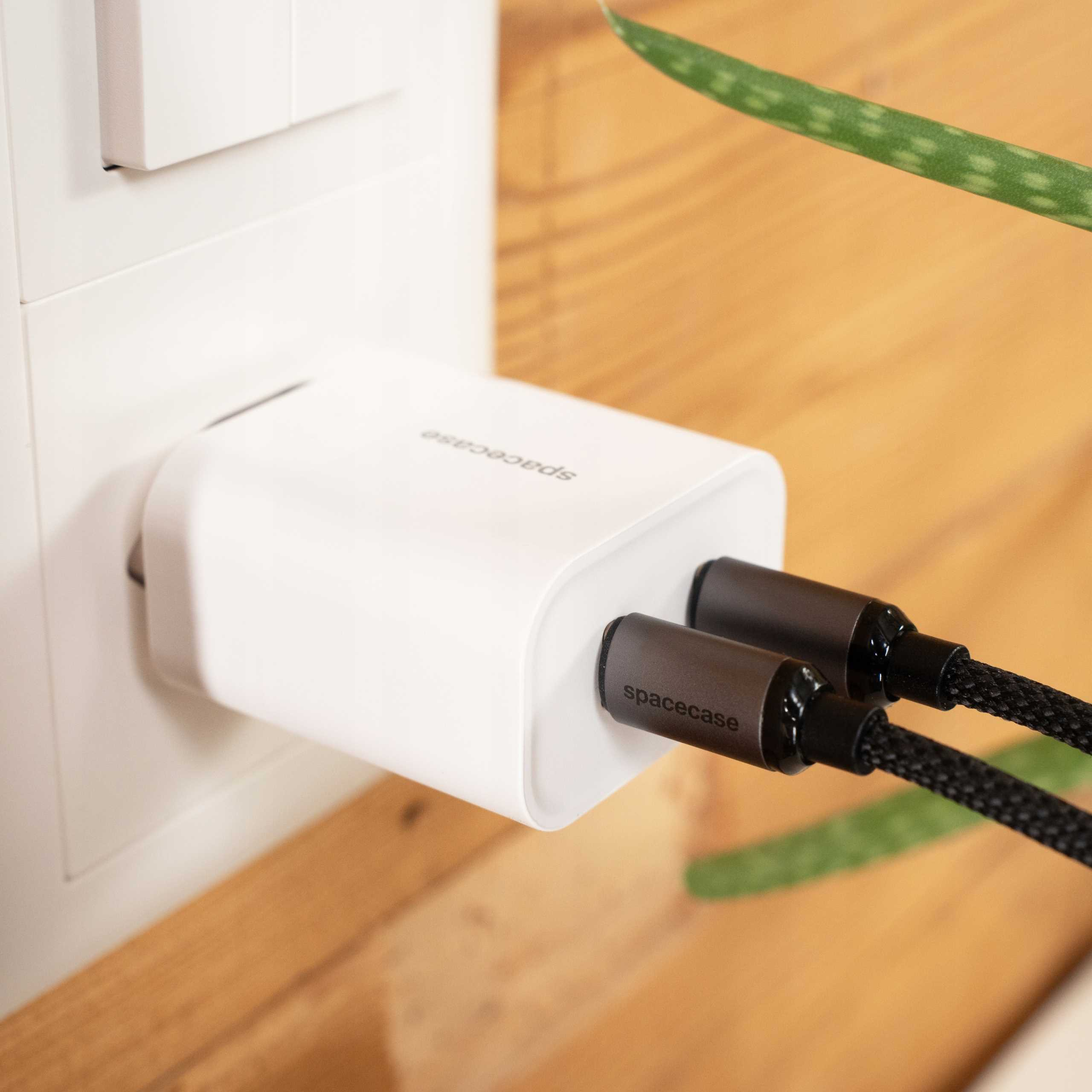 Biała ładowarka z dwoma czarnymi kablami włożonymi do gniazdka. Zielona roślina w tle.