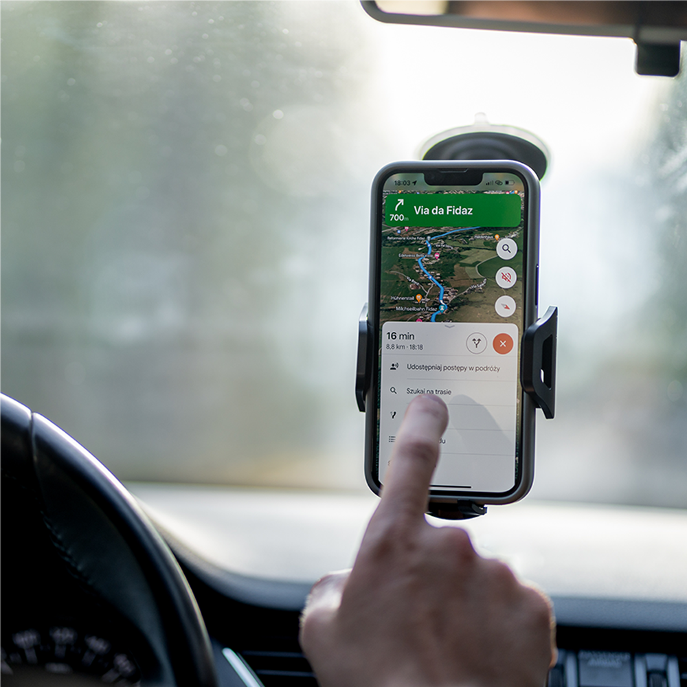 Telefon wyświetla aplikację nawigacji map. Jest zamontowany na przedniej szybie samochodu. Palec dotyka ekranu. Kierownica jest na pierwszym planie.