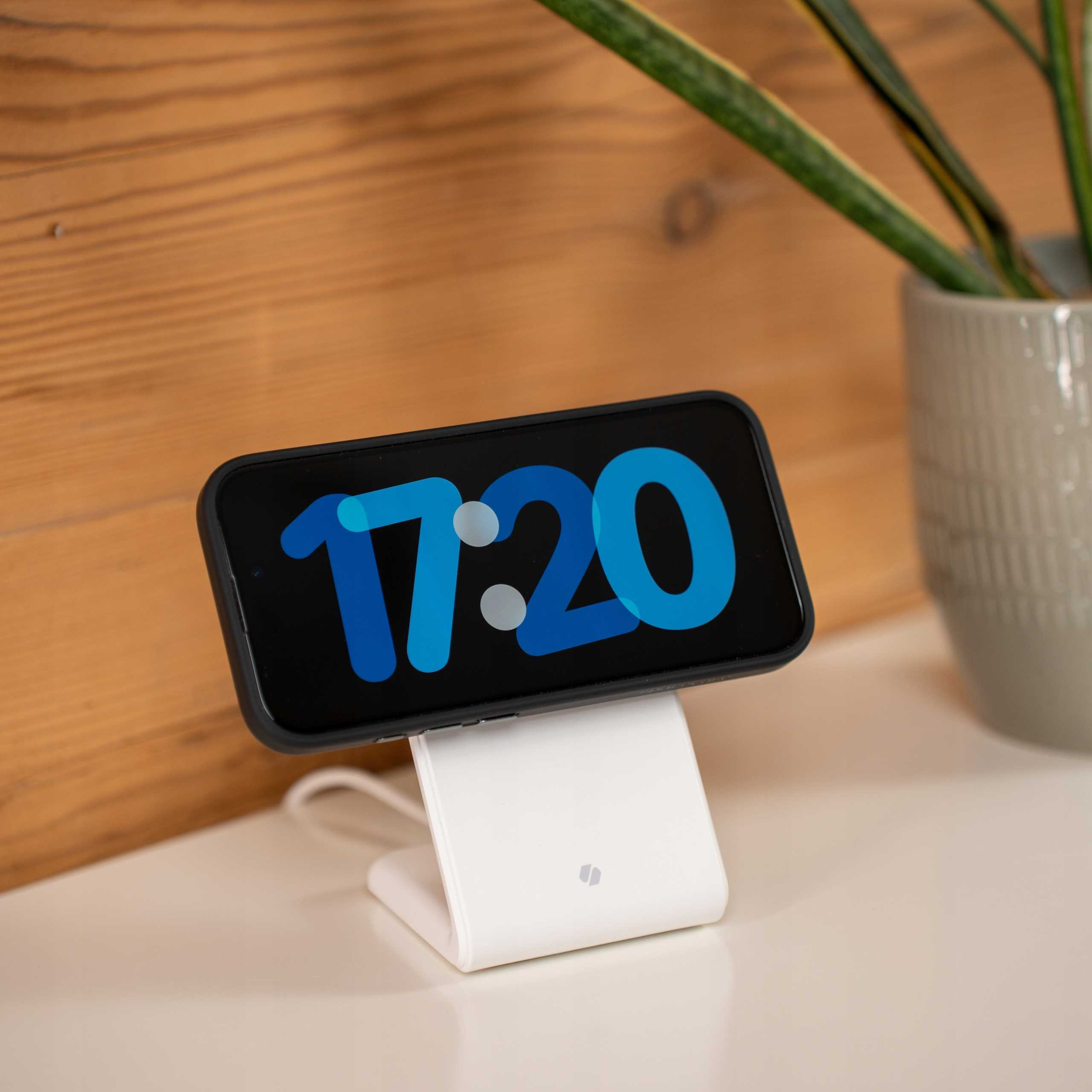Telefon wyświetla godzinę 17:20, ładując się na białej podstawce. Drewniane tło, roślina w szarej donicy.
