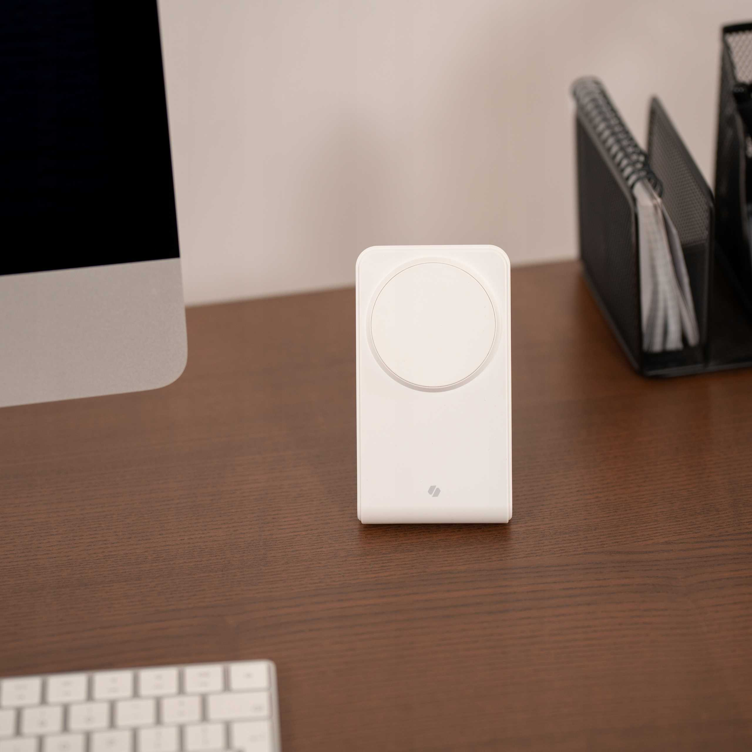 Biała ładowarka bezprzewodowa na drewnianym biurku. Widoczny jest również ekran komputera, klawiatura i czarny organizer.