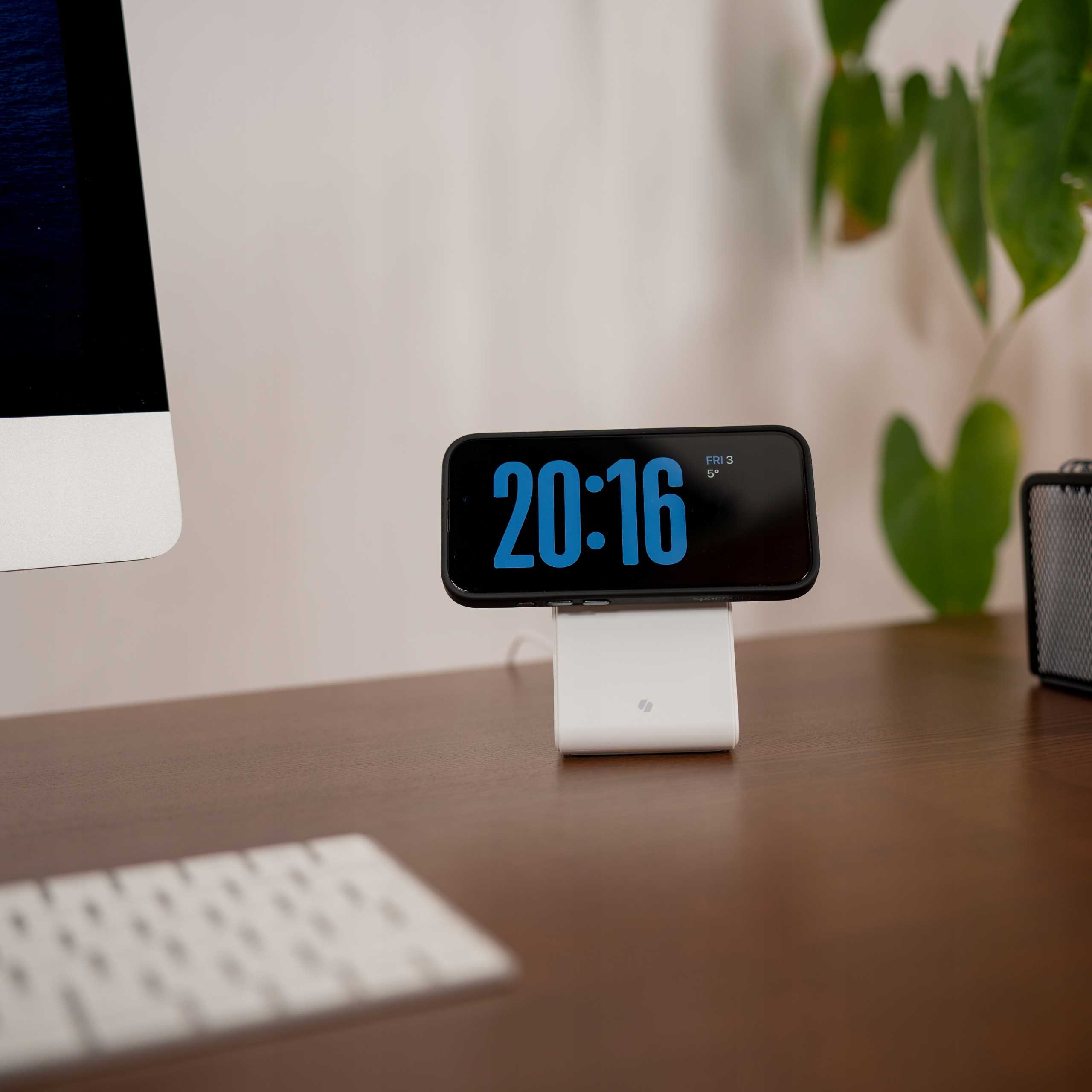 Telefon z godziną 20:16 spoczywa na białej ładowarce. Klawiatura i komputer w tle.