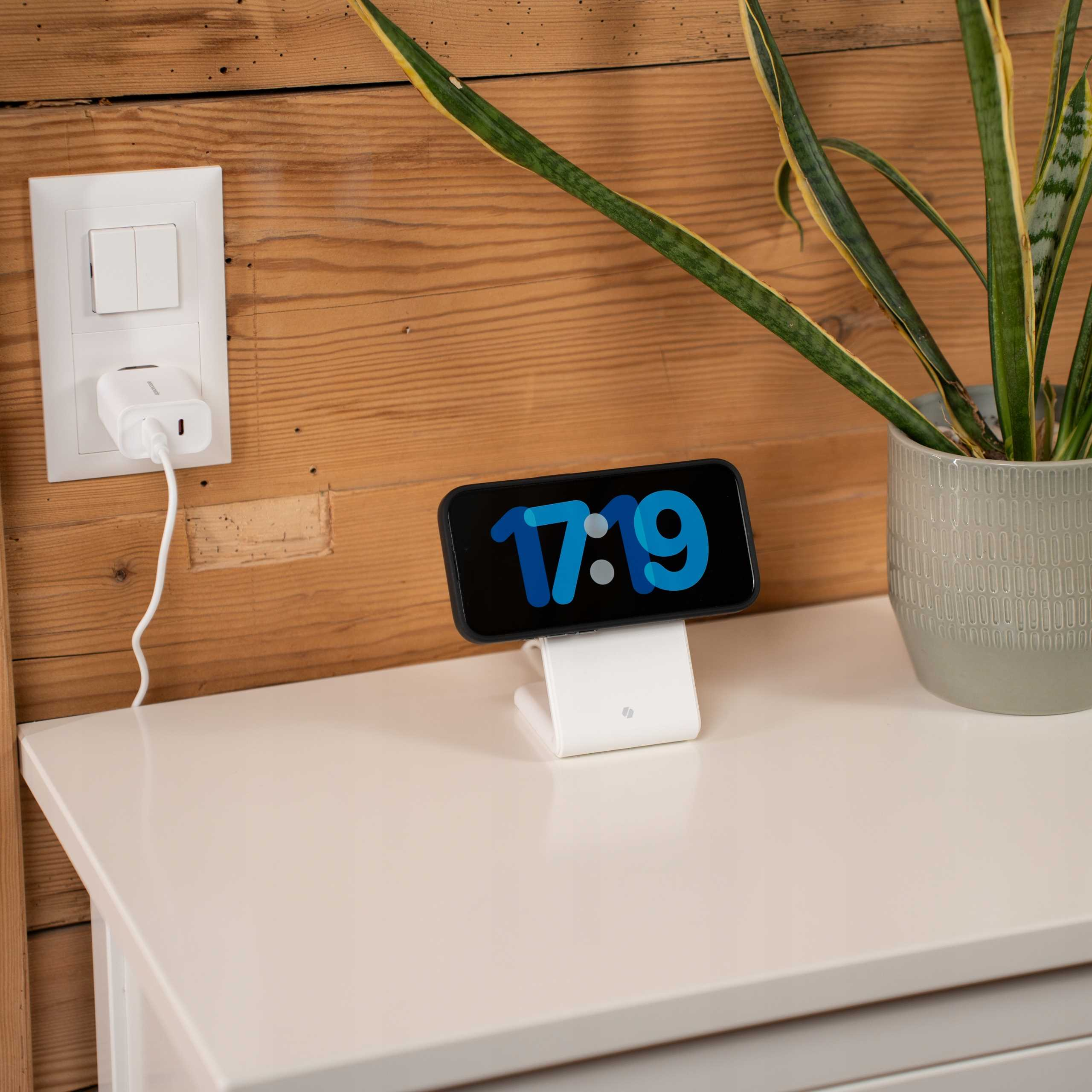 Telefon z godziną 17:19 spoczywa na białym stojaku. Po prawej roślina, po lewej gniazdko.