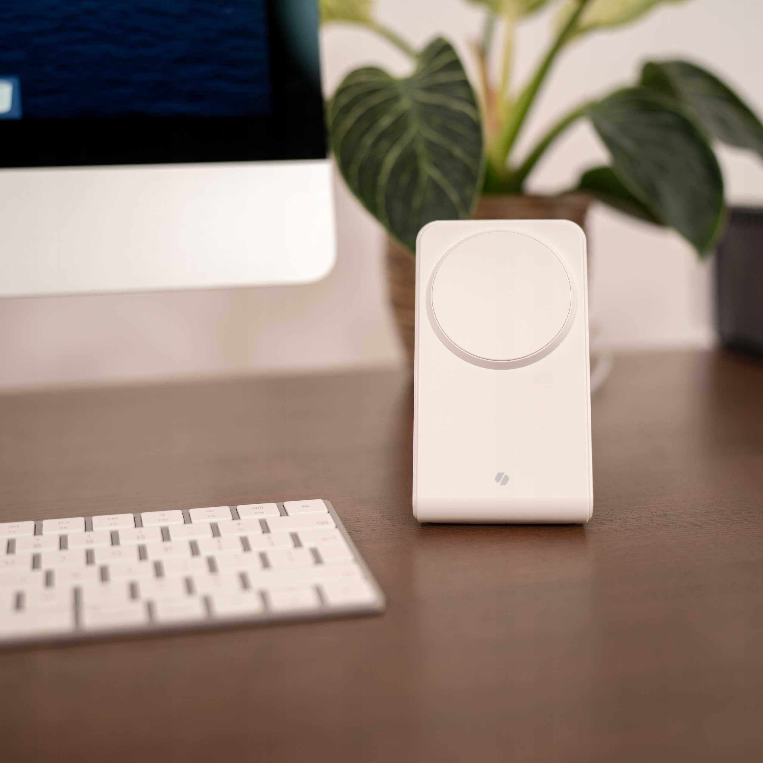 Biała ładowarka bezprzewodowa stoi na brązowym biurku. W tle widoczna klawiatura i roślina.