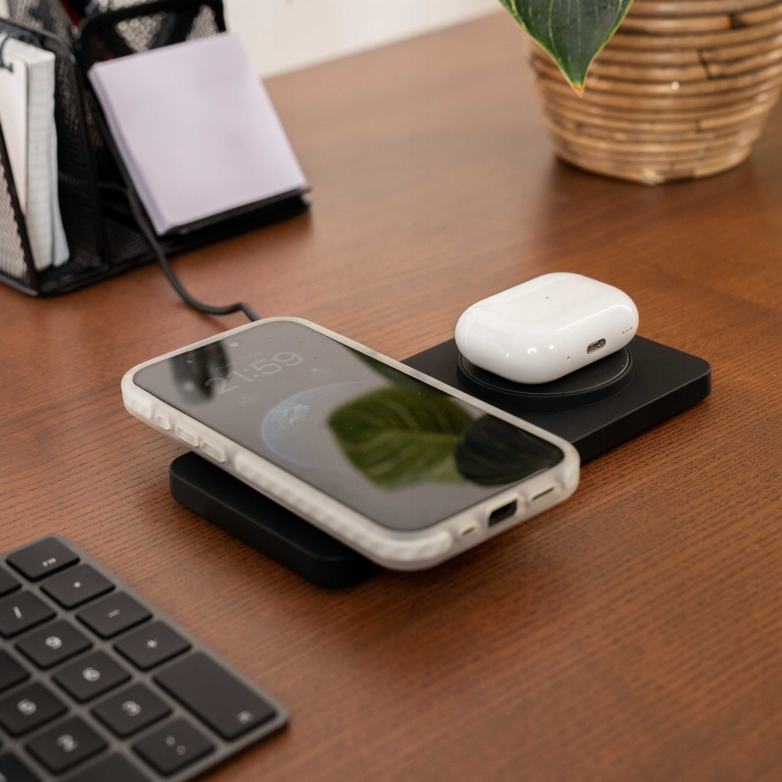 Telefon i słuchawki ładują się na ładowarce bezprzewodowej. Widoczna jest również klawiatura, roślina i notatnik.
