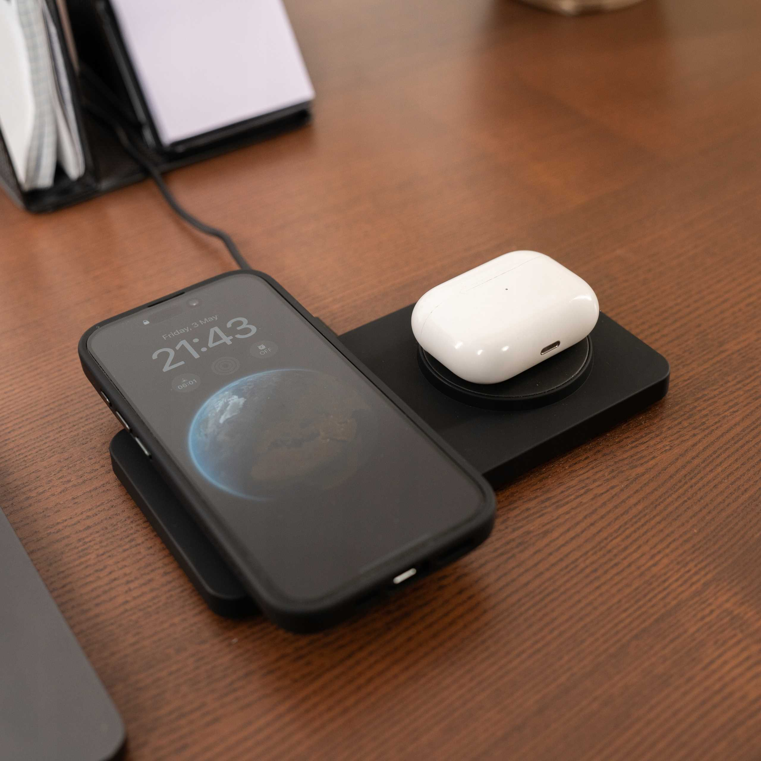 Czarny telefon na ładowarce. Białe AirPods na ładowarce. Drewniany stół. Czas: 21:43.