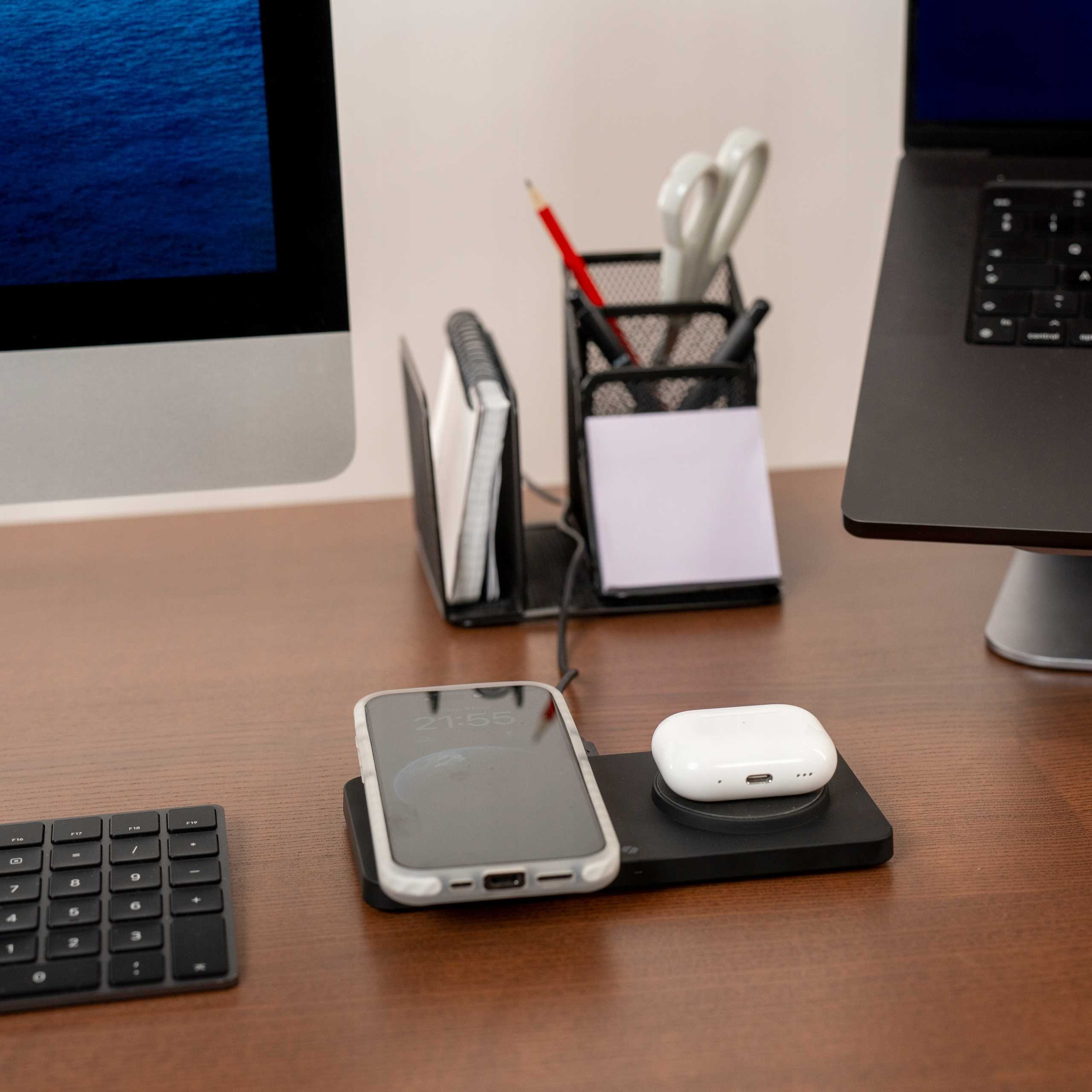Konfiguracja biurka z ładowaniem telefonu, AirPods, klawiaturą i monitorem. Widoczny jest pojemnik na długopisy z długopisami i nożyczkami.