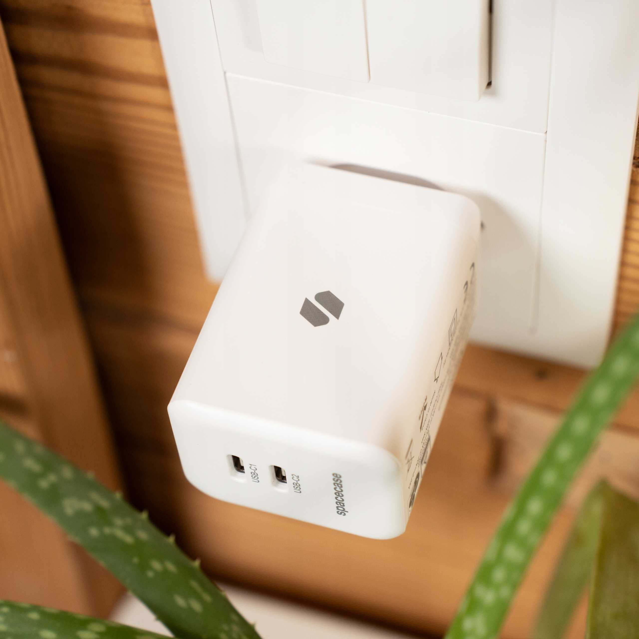 Biała ładowarka USB jest podłączona do gniazdka ściennego. Zielona roślina jest na pierwszym planie. Ściana jest biała.