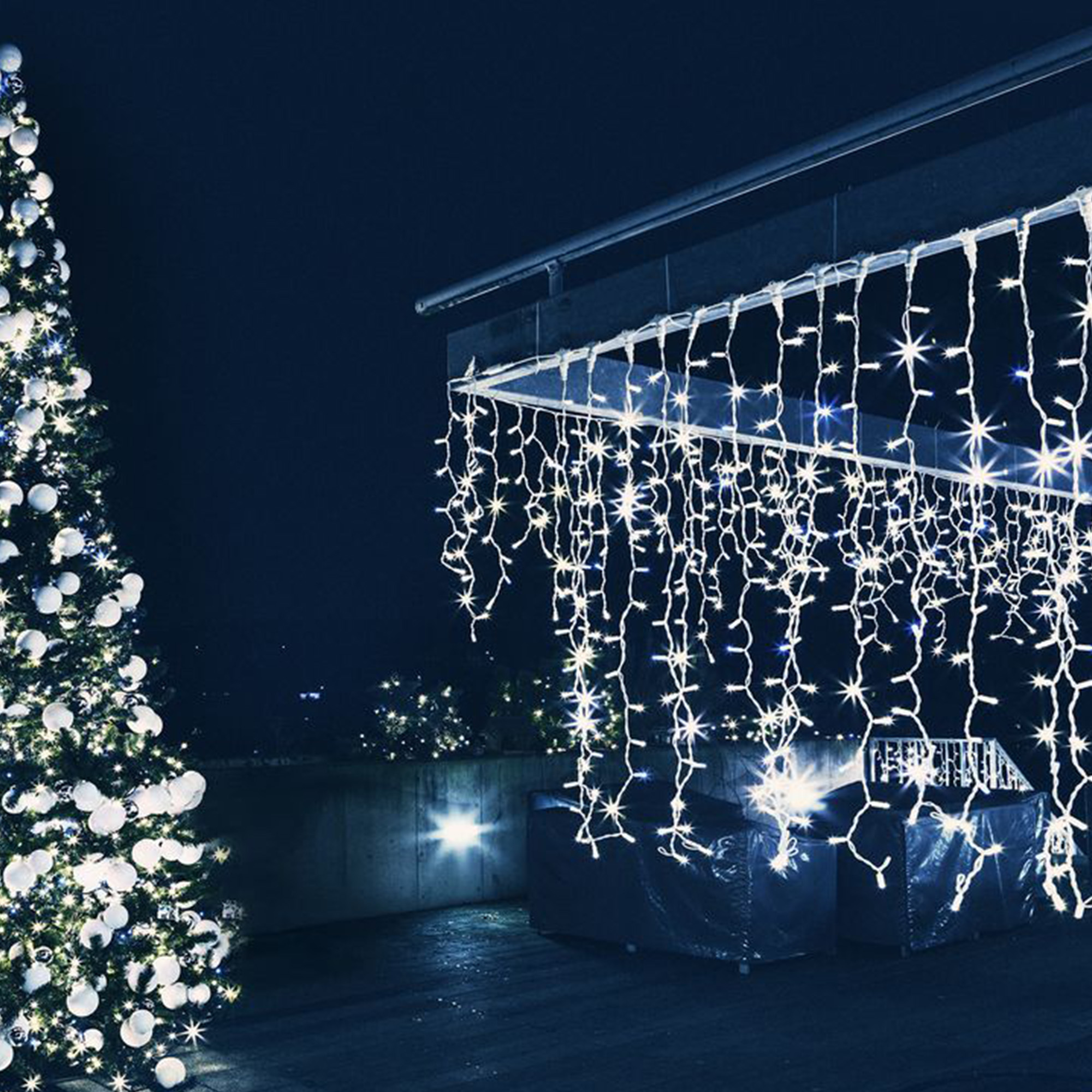 Un árbol de Navidad con adornos y luces blancas. Luces de cadena blancas cuelgan de una estructura. El cielo nocturno es azul oscuro.