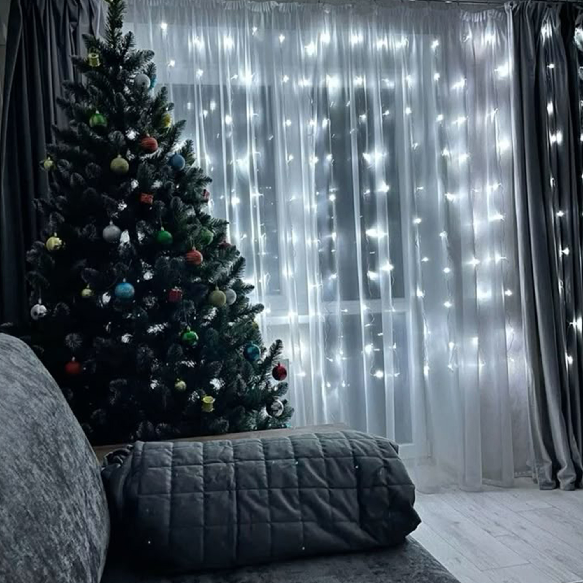 Un árbol de Navidad decorado está a la izquierda. Luces detrás de cortinas transparentes y cortinas oscuras enmarcan el árbol. Se ve un sofá y una almohada.