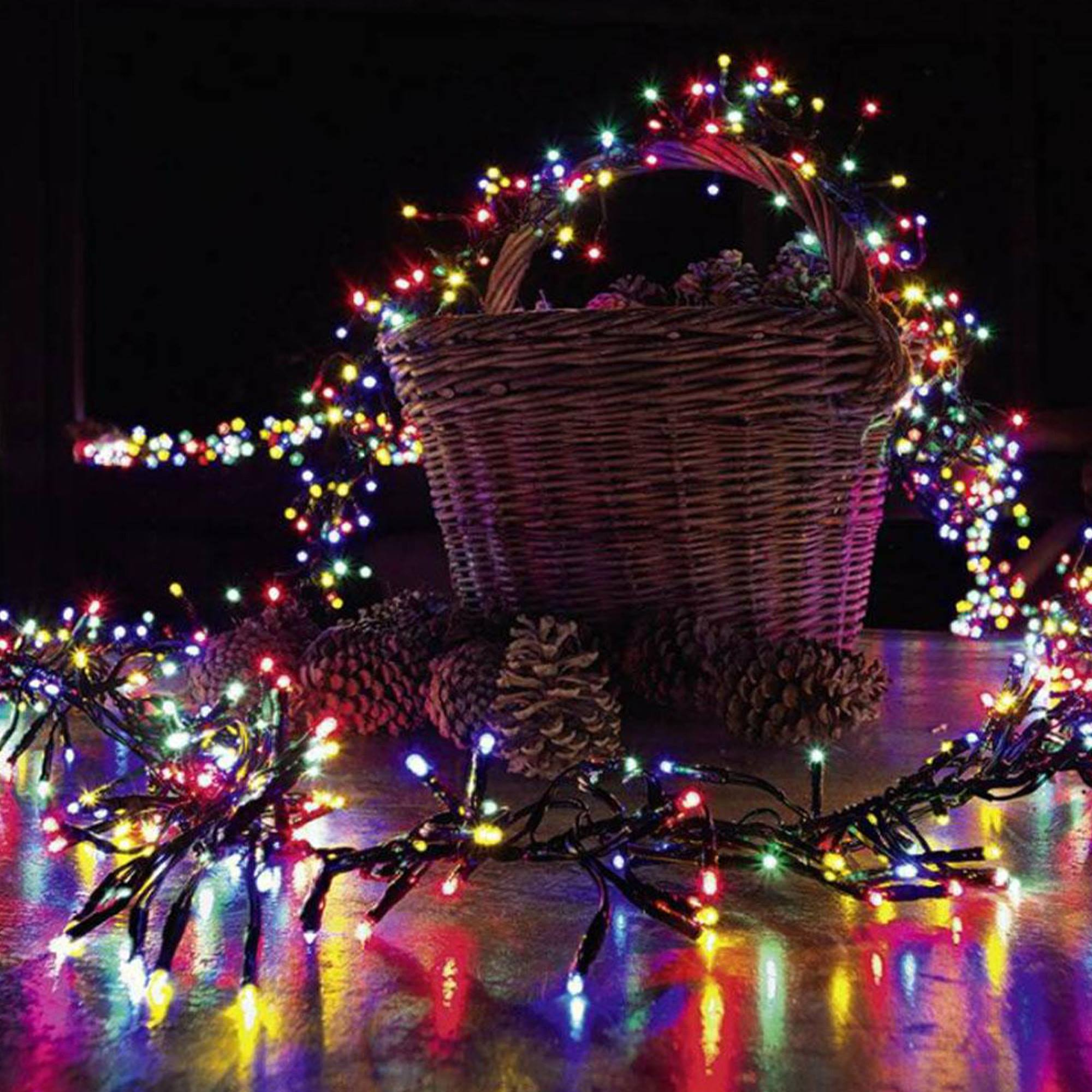 Una cesta de mimbre con piñas y luces de colores. Las luces son rojas, azules, verdes y amarillas. Fondo oscuro.