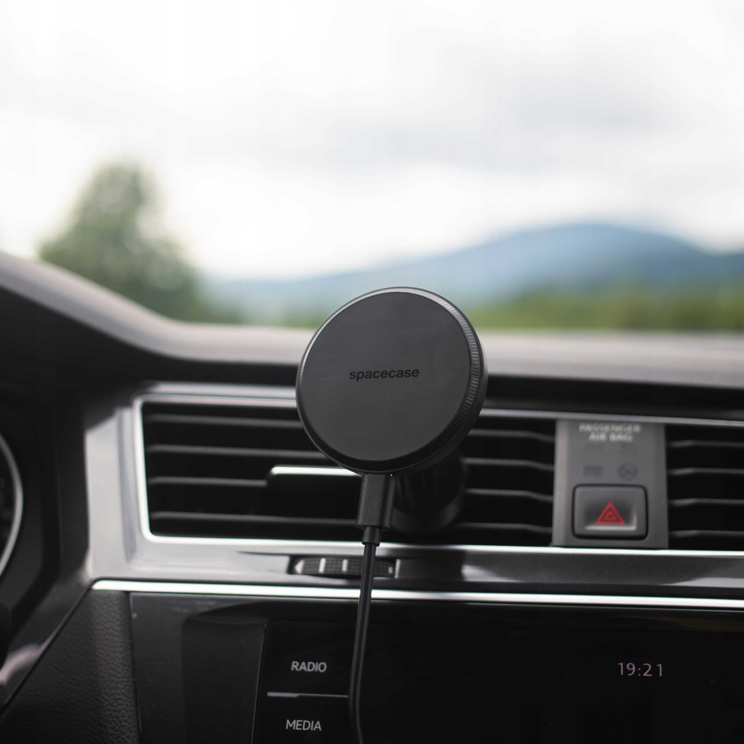 Czarny uchwyt na telefon z napisem 'spacecase' jest przymocowany do deski rozdzielczej samochodu. W tle widoczne są góry.