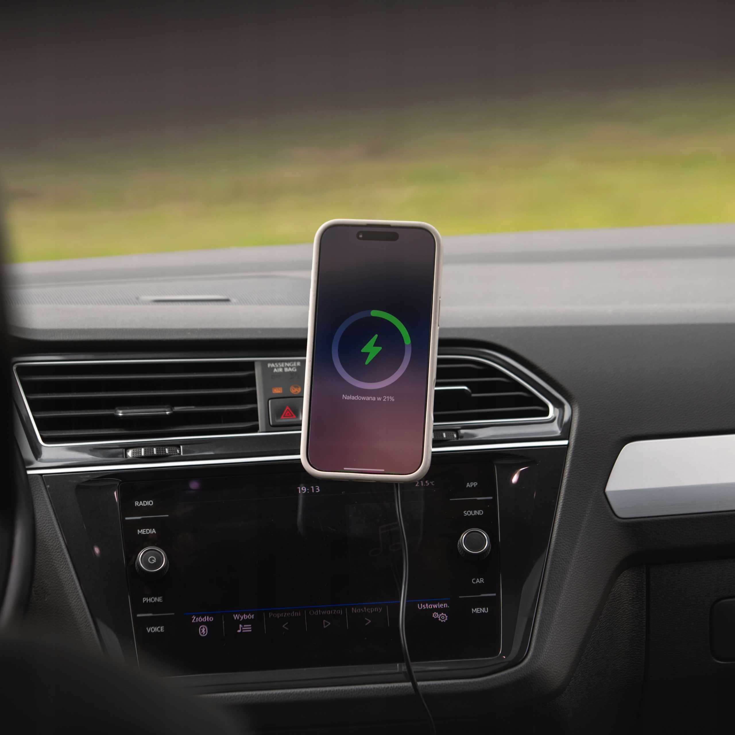 Telefon ładuje się w samochodzie. Ekran pokazuje 21% naładowania, z zieloną błyskawicą. Deska rozdzielcza jest czarna.