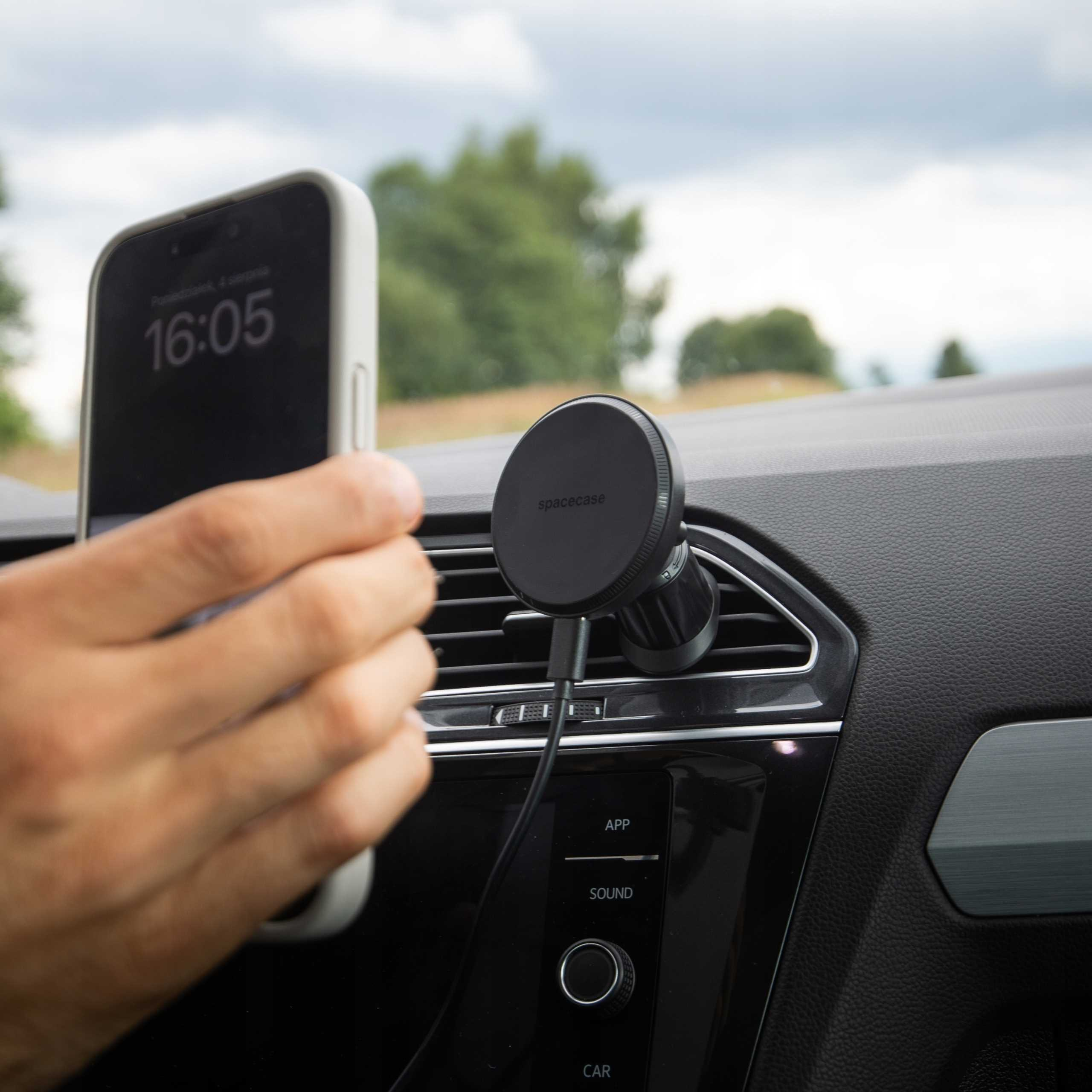 Osoba umieszcza telefon w uchwycie samochodowym. Telefon wyświetla godzinę 16:05. Uchwyt jest przymocowany do kratki wentylacyjnej.