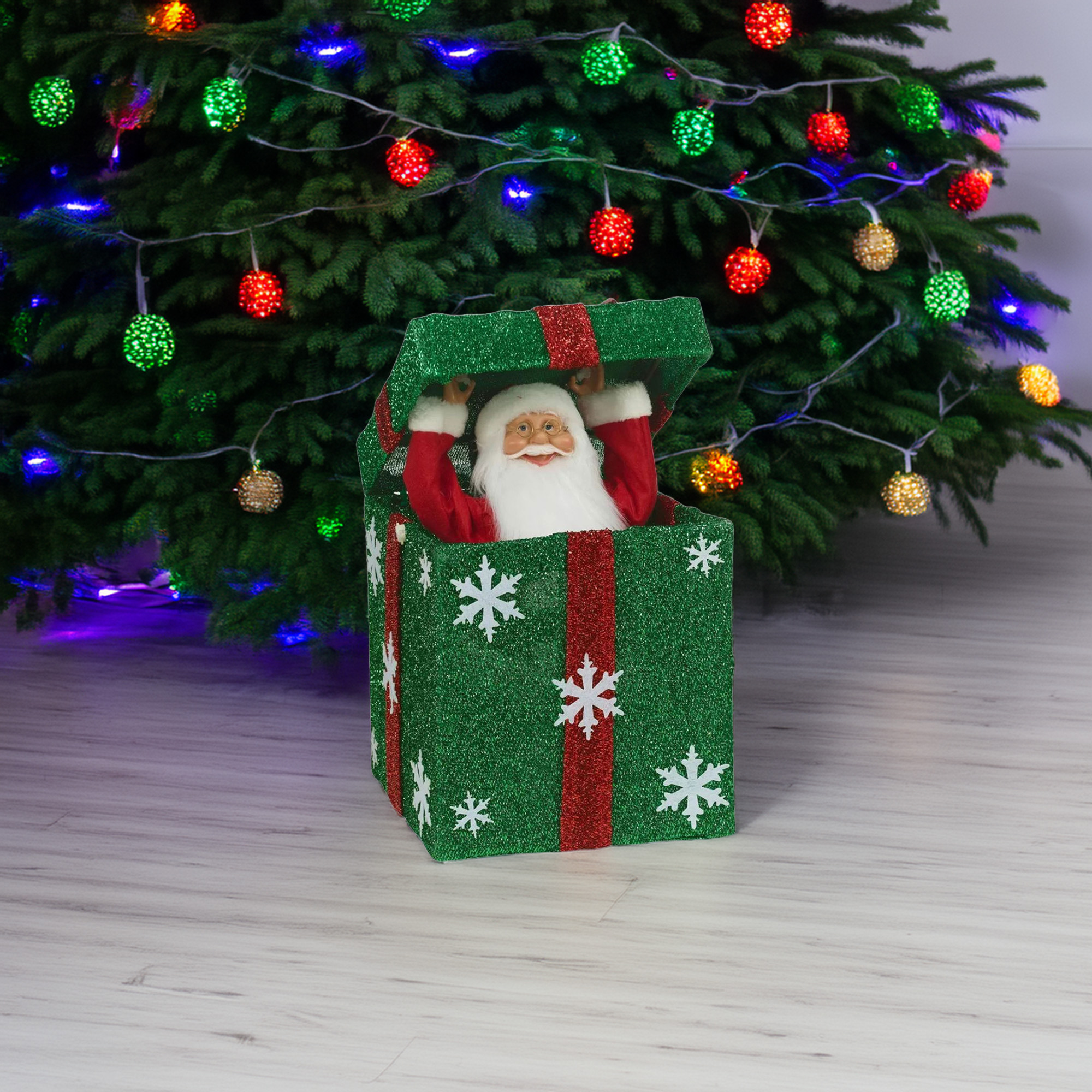 Figura de Papá Noel en una caja de regalo verde con copos de nieve, frente a un árbol de Navidad decorado con luces.