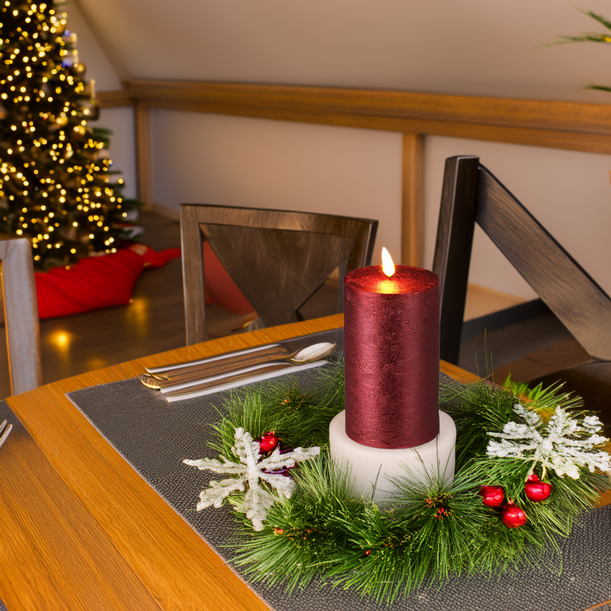 Una vela roja encendida está en un centro de mesa navideño con adornos, copos de nieve y un árbol al fondo.