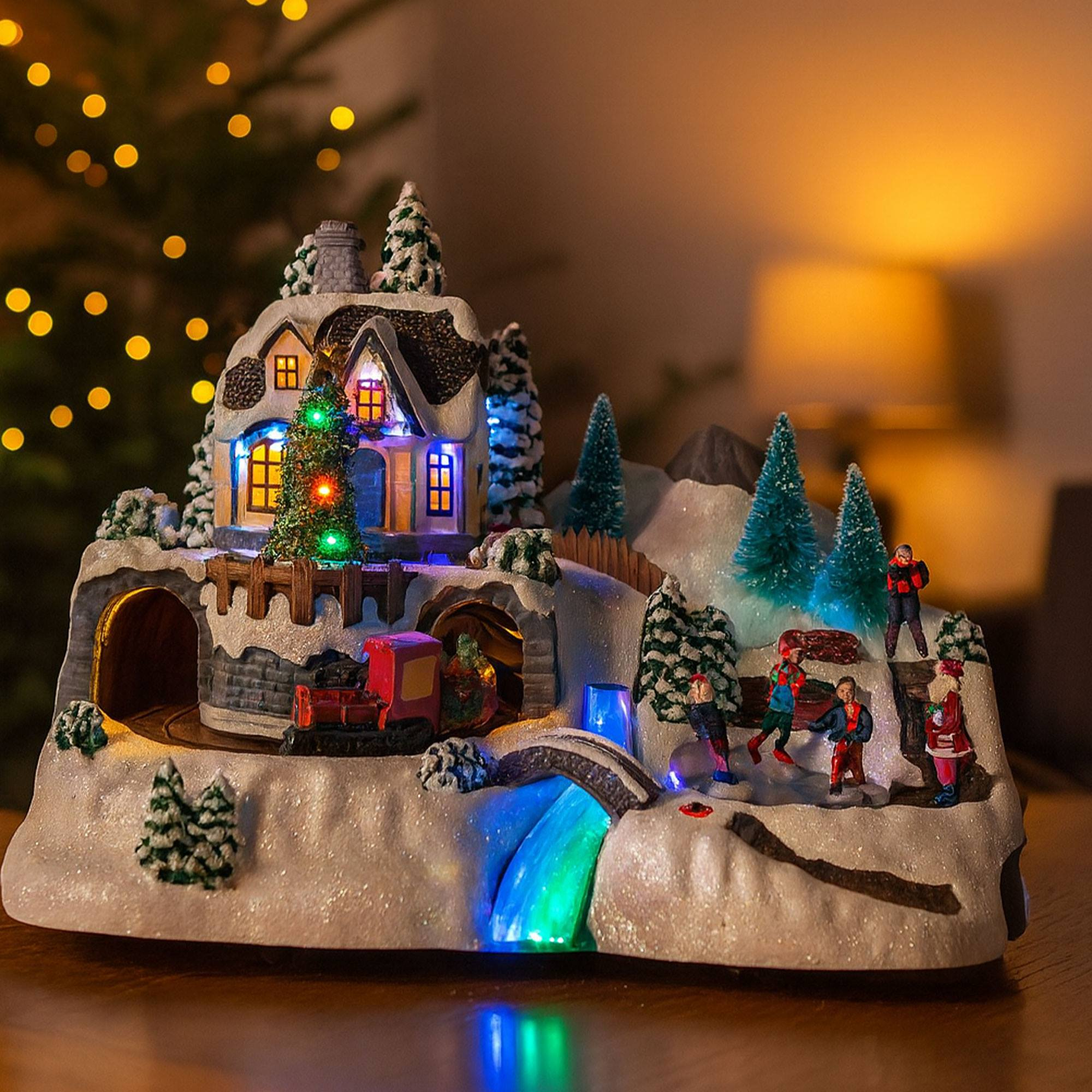 Una escena navideña en miniatura con una casa, tren, figuras, árboles y una cascada. Las luces iluminan la escena.