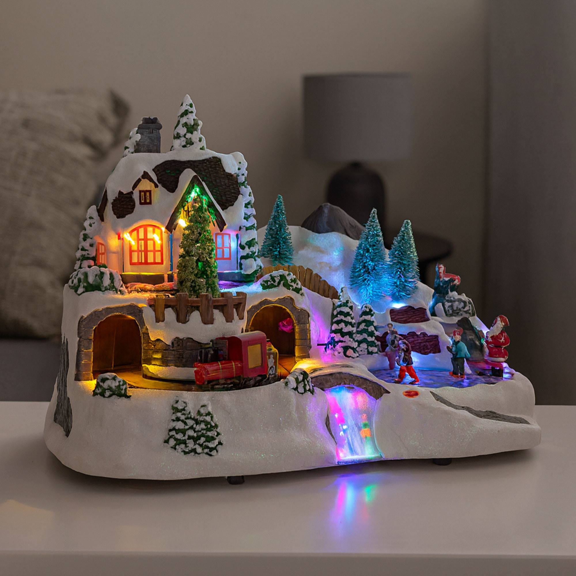 Una escena navideña con una casa, un tren, gente patinando sobre hielo y Papá Noel. Las luces brillan. La nieve cubre la escena.