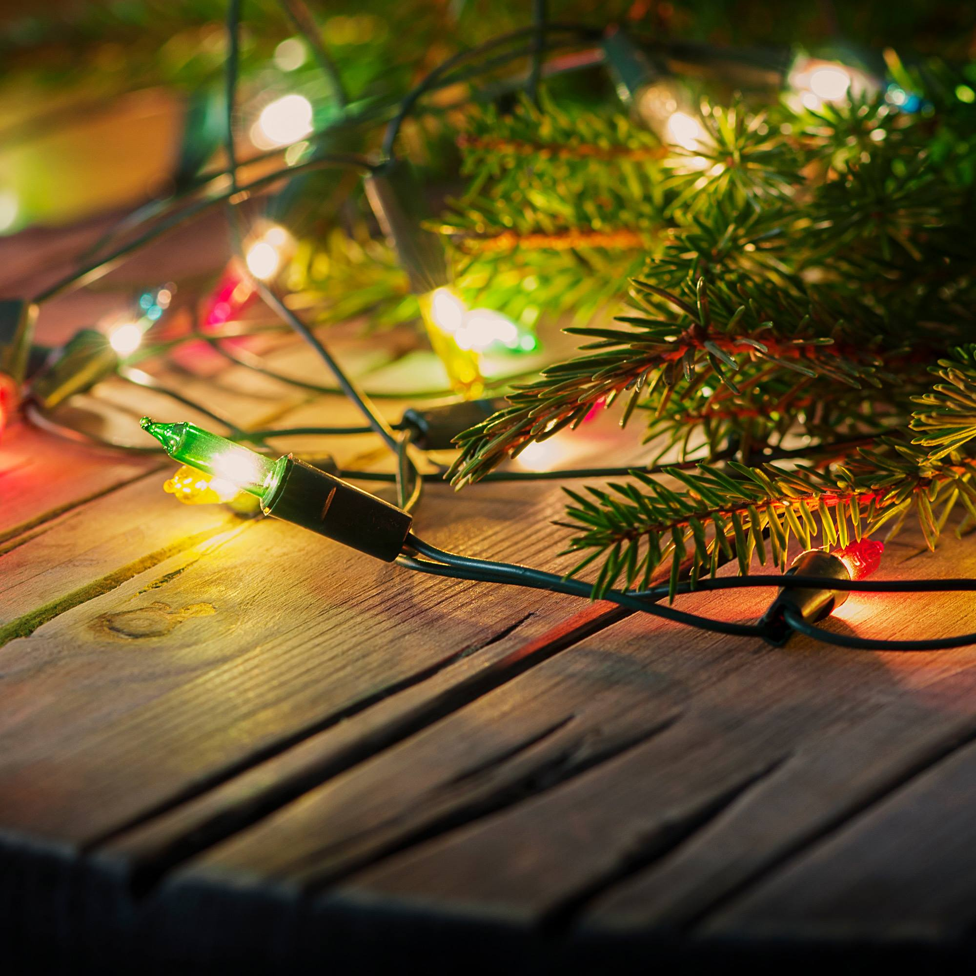 Primer plano de luces navideñas con una bombilla verde enfocada, sobre madera con una rama de abeto.