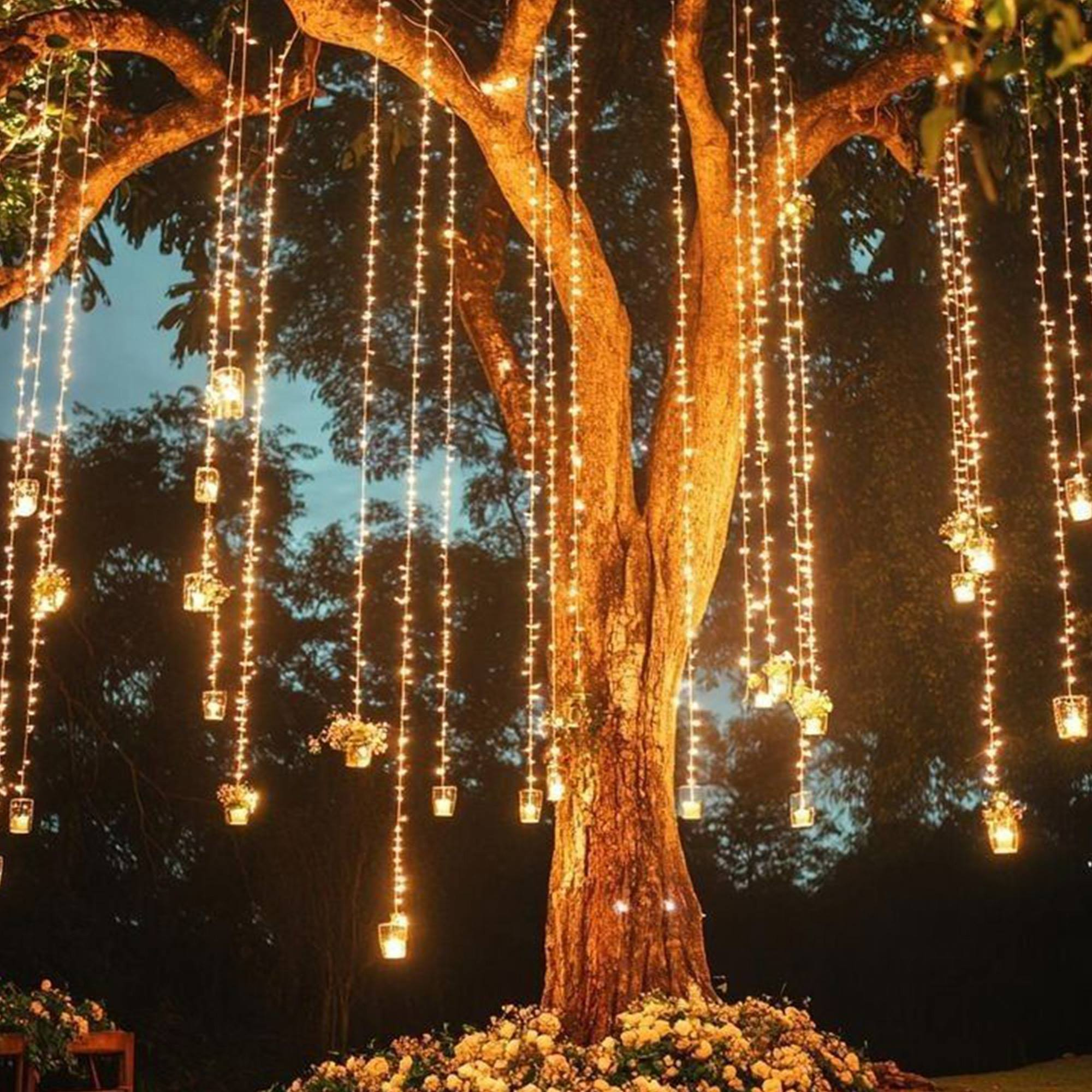 Un árbol decorado con luces colgantes y pequeños frascos con velas. Flores en la base.