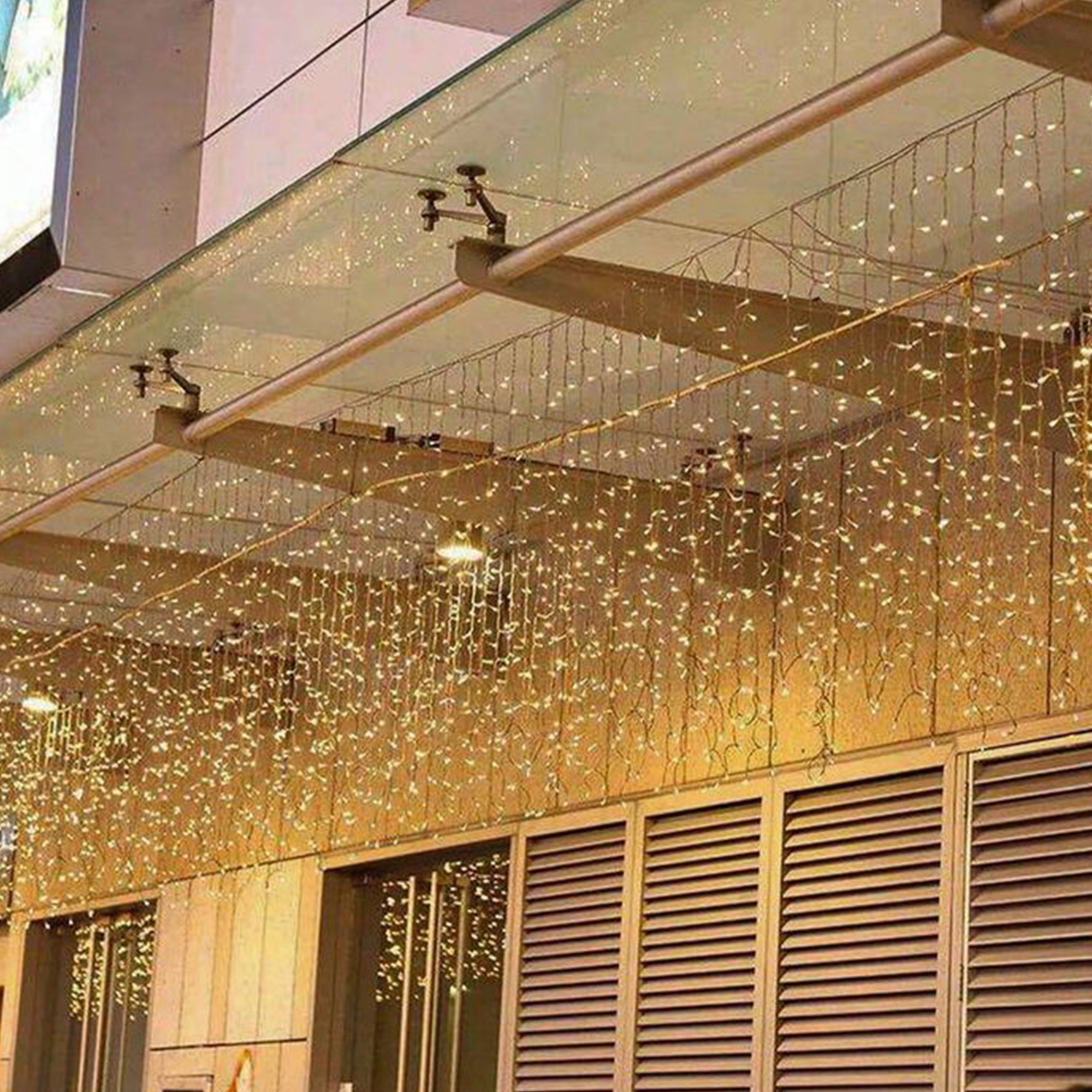 Vista exterior con hilos de pequeñas luces colgando de un techo de vidrio. Debajo, hay ventanas y paneles de madera.