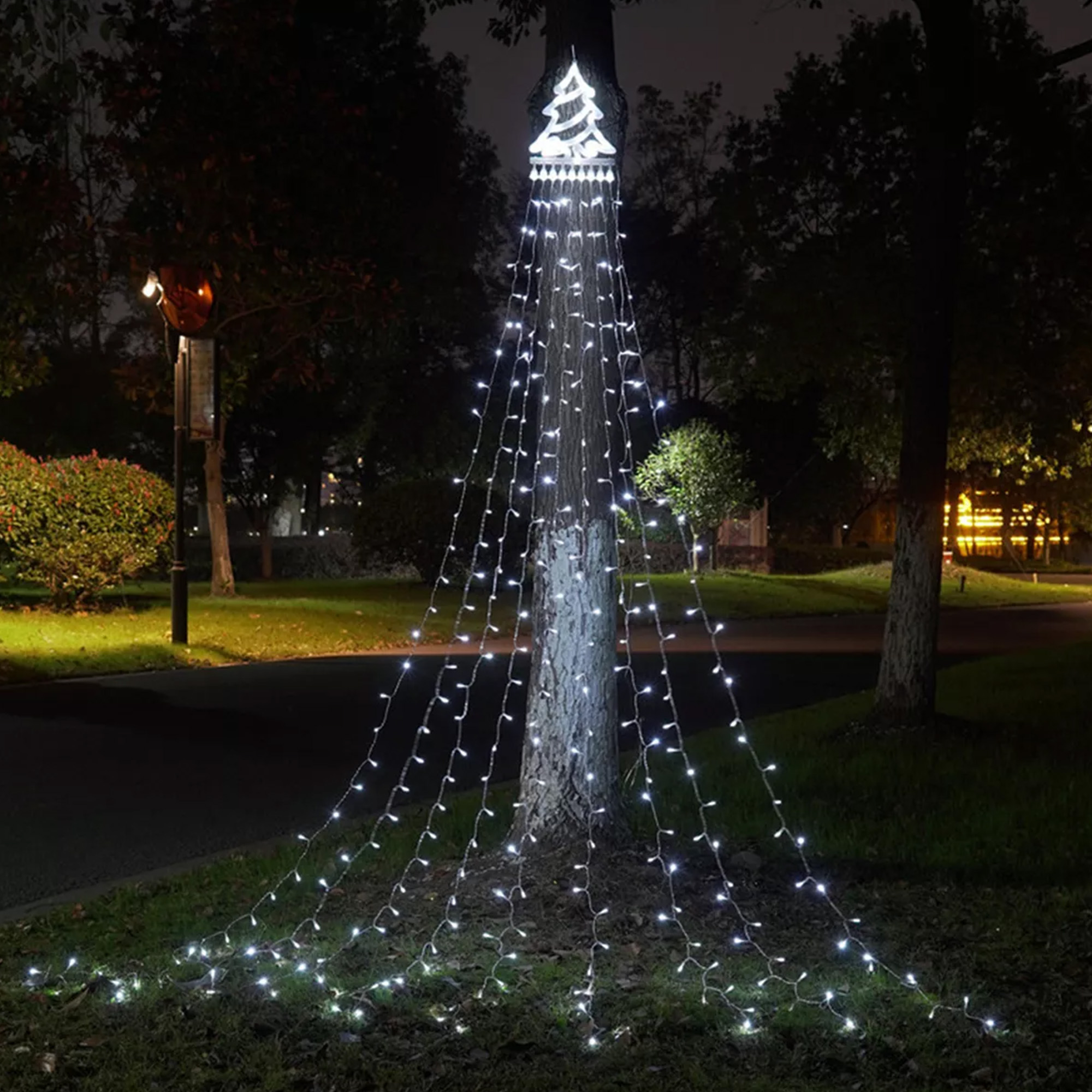 Un árbol decorado con luces navideñas blancas en forma de árbol, por la noche. Se ven otros árboles y luces del parque.