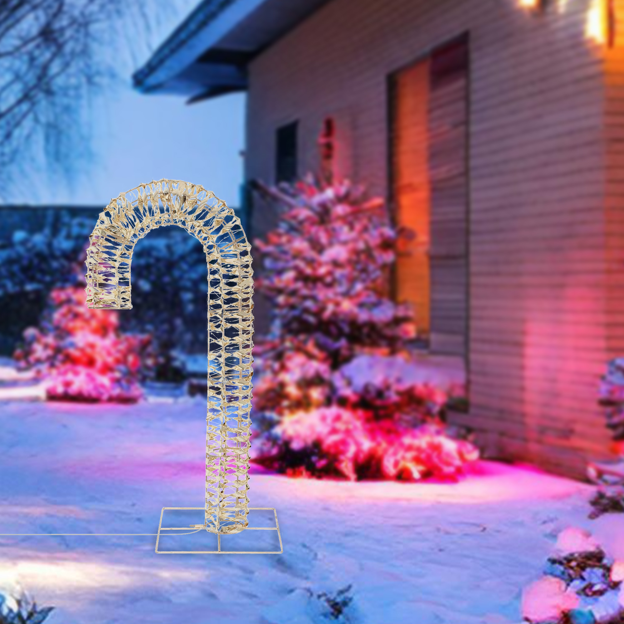 Una gran decoración dorada de bastón de caramelo está en un jardín nevado. Hay árboles y una casa iluminada en el fondo.