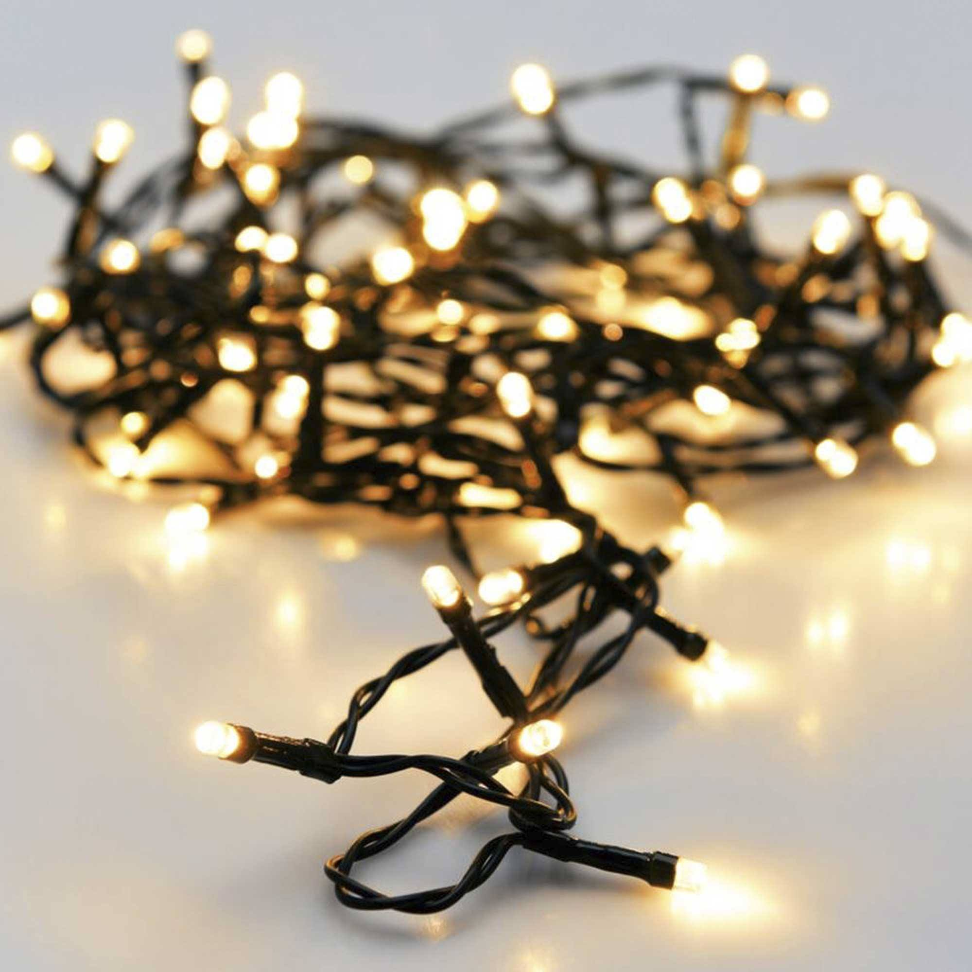 Luces de cadena con pequeños LED blancos cálidos en un cable negro, sobre una superficie blanca. Algunas luces están enfocadas.