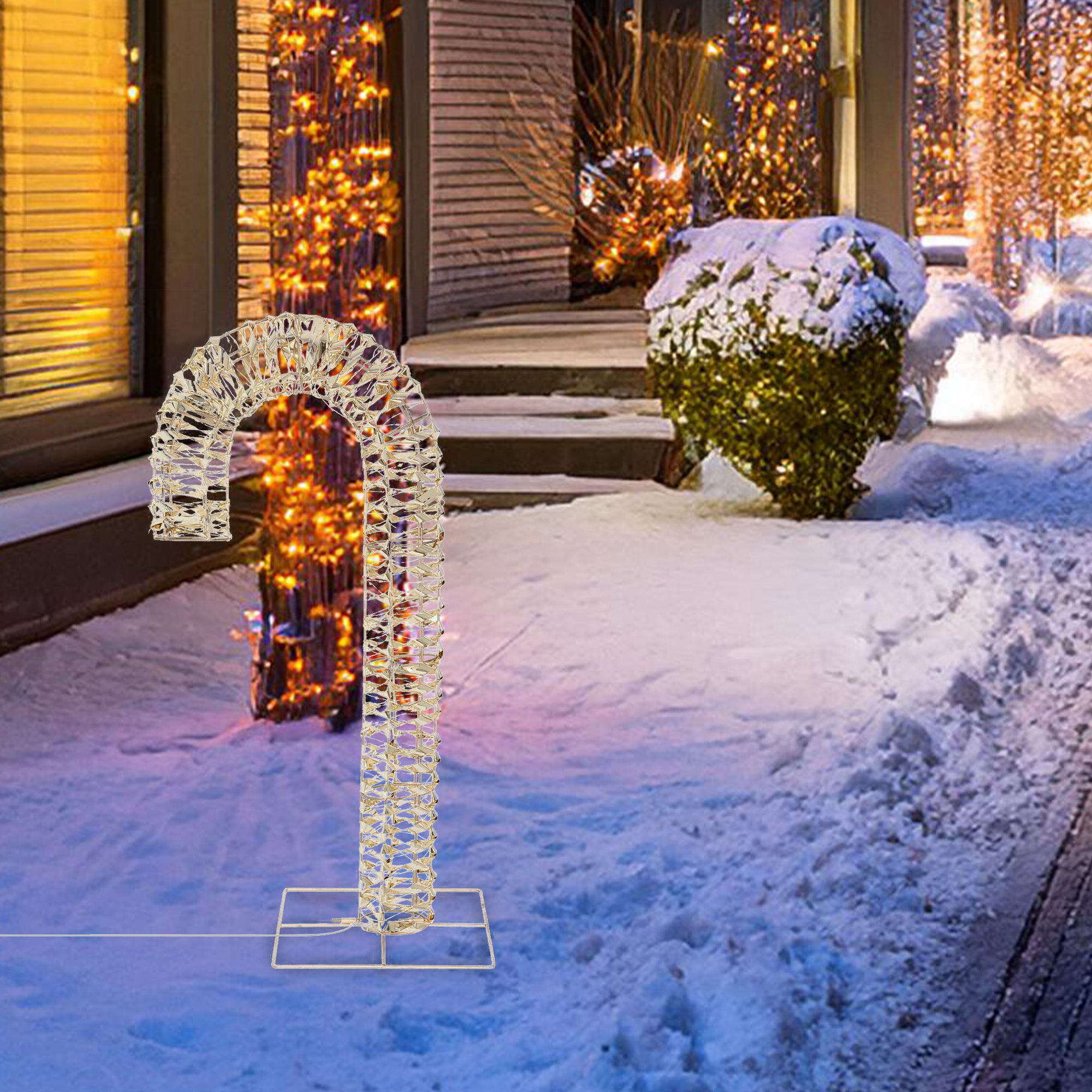 Un bastón de caramelo decorativo iluminado en la nieve. Luces adornan la casa y arbustos en el fondo. Invierno.