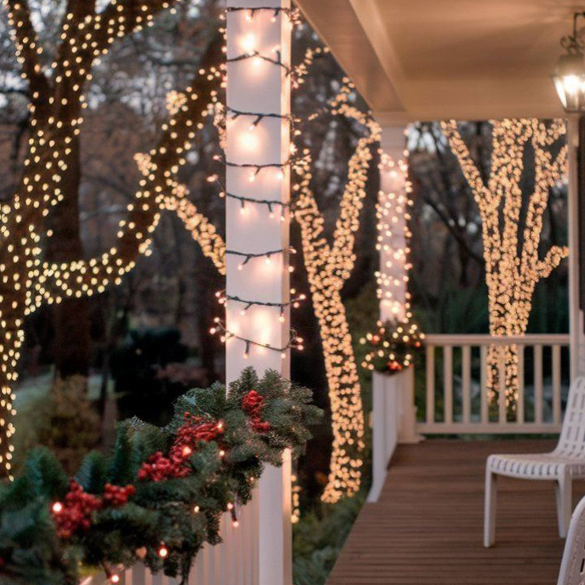 Un porche decorado con luces navideñas y guirnaldas. Los árboles están iluminados con luces. Sillas blancas y barandillas del porche.