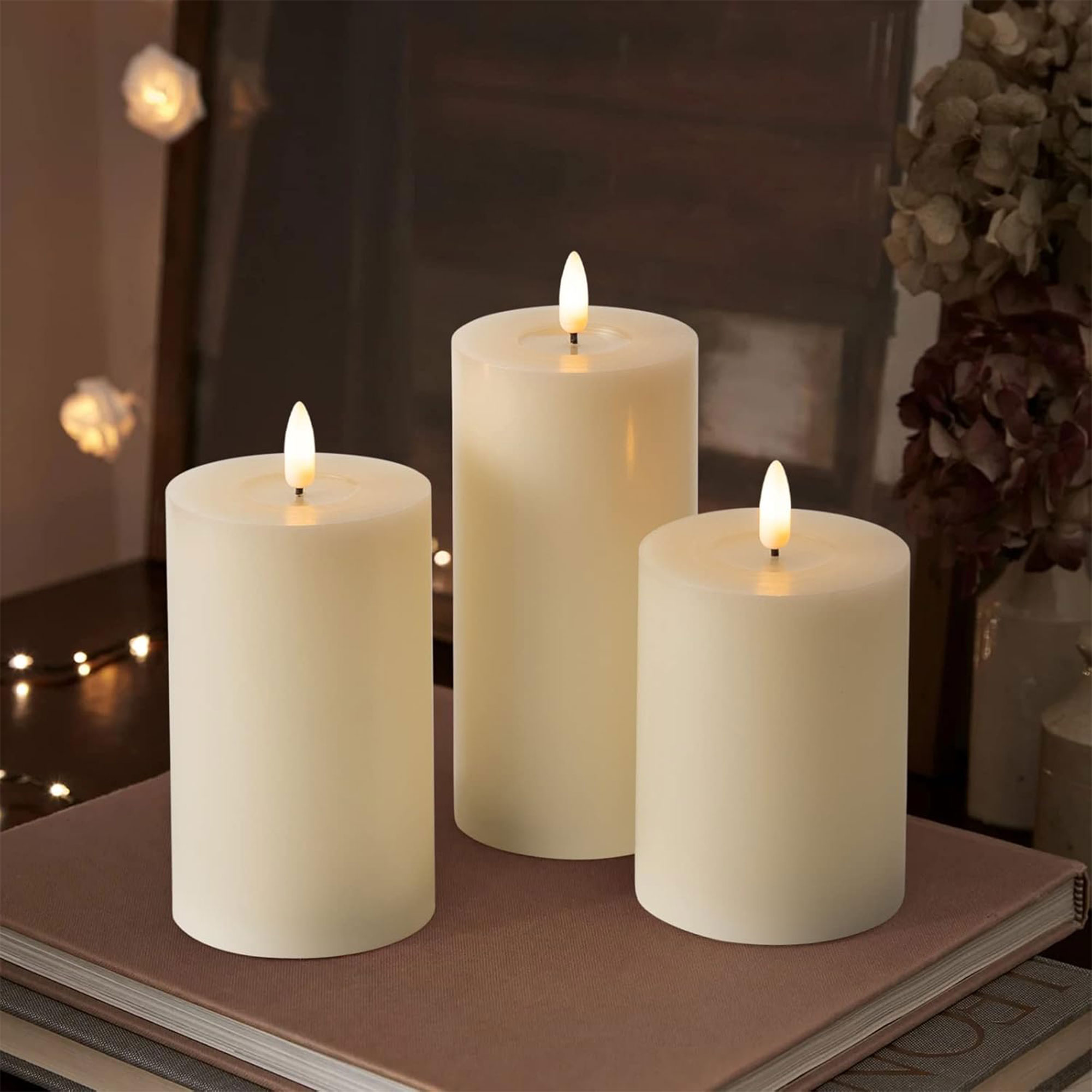 Tres velas marfil con llamas encendidas sobre libros. El fondo incluye flores y luces.
