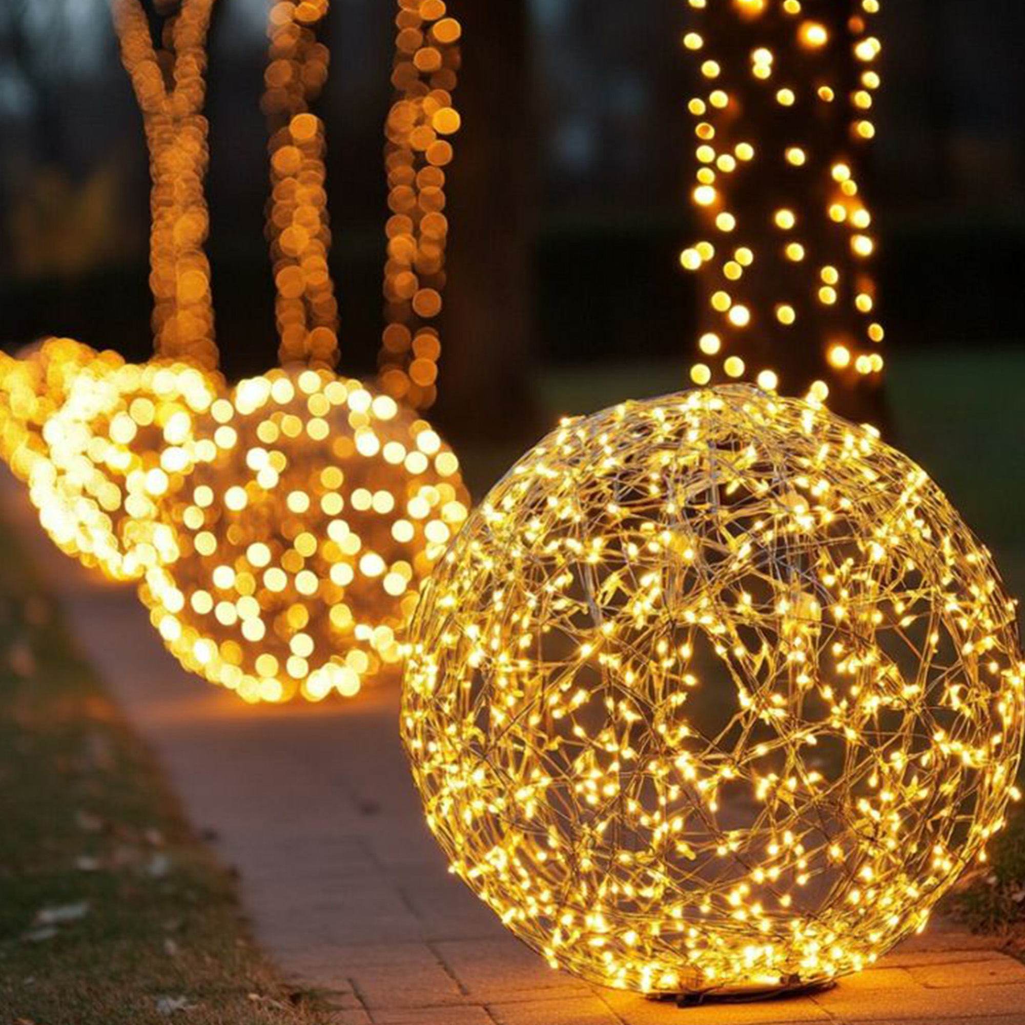 Luces esféricas brillantes en un camino, con troncos de árboles adornados con luces al fondo. Escena nocturna.