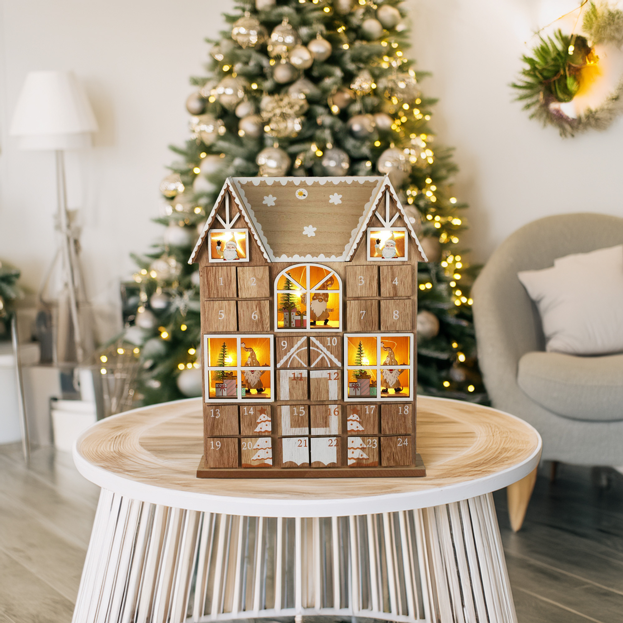 Un calendario de adviento de madera con forma de casa está sobre una mesa. Un árbol de Navidad está detrás. Otras decoraciones.