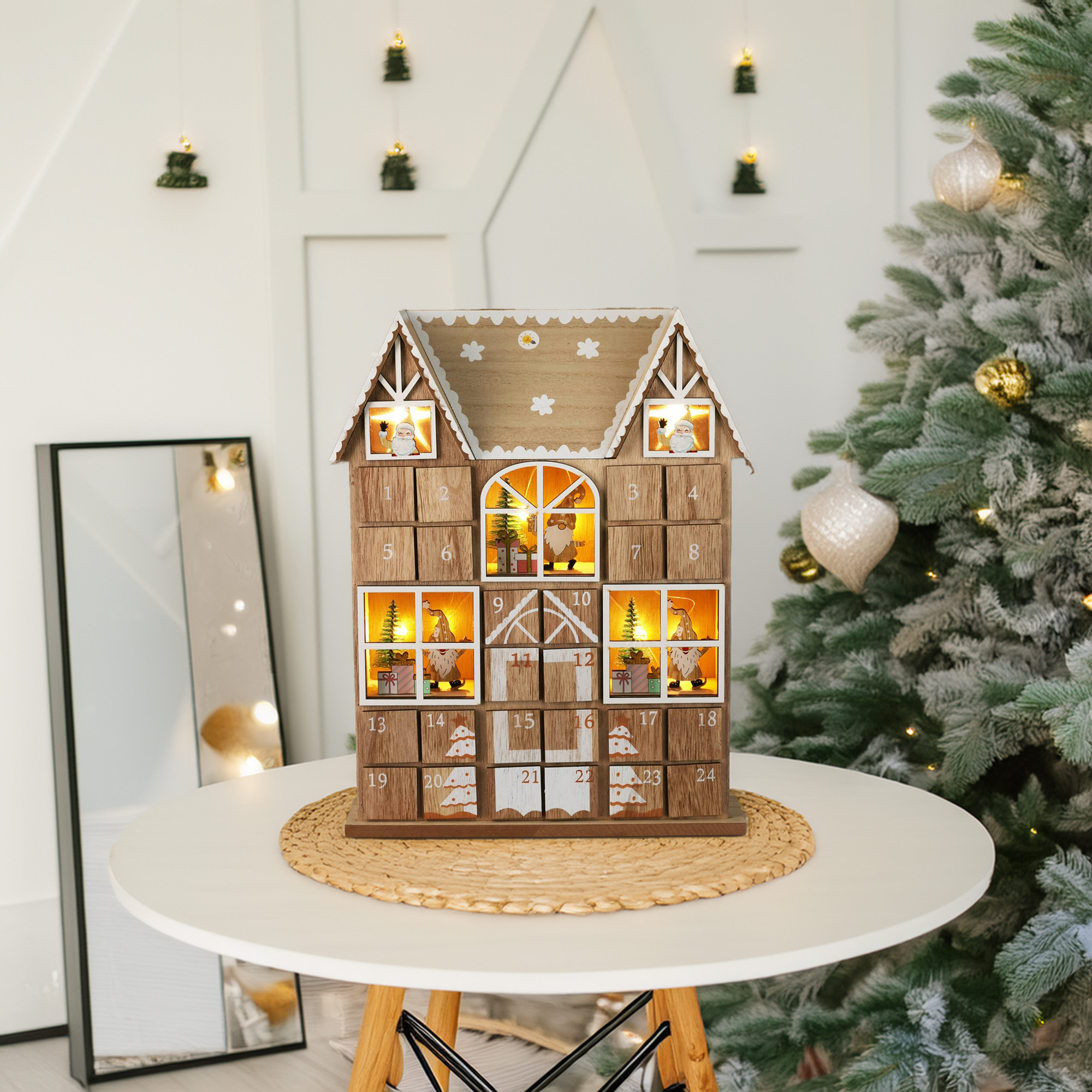 Un calendario de adviento de madera con forma de casa, sobre una mesa blanca con un árbol de Navidad a la derecha. El fondo es blanco.