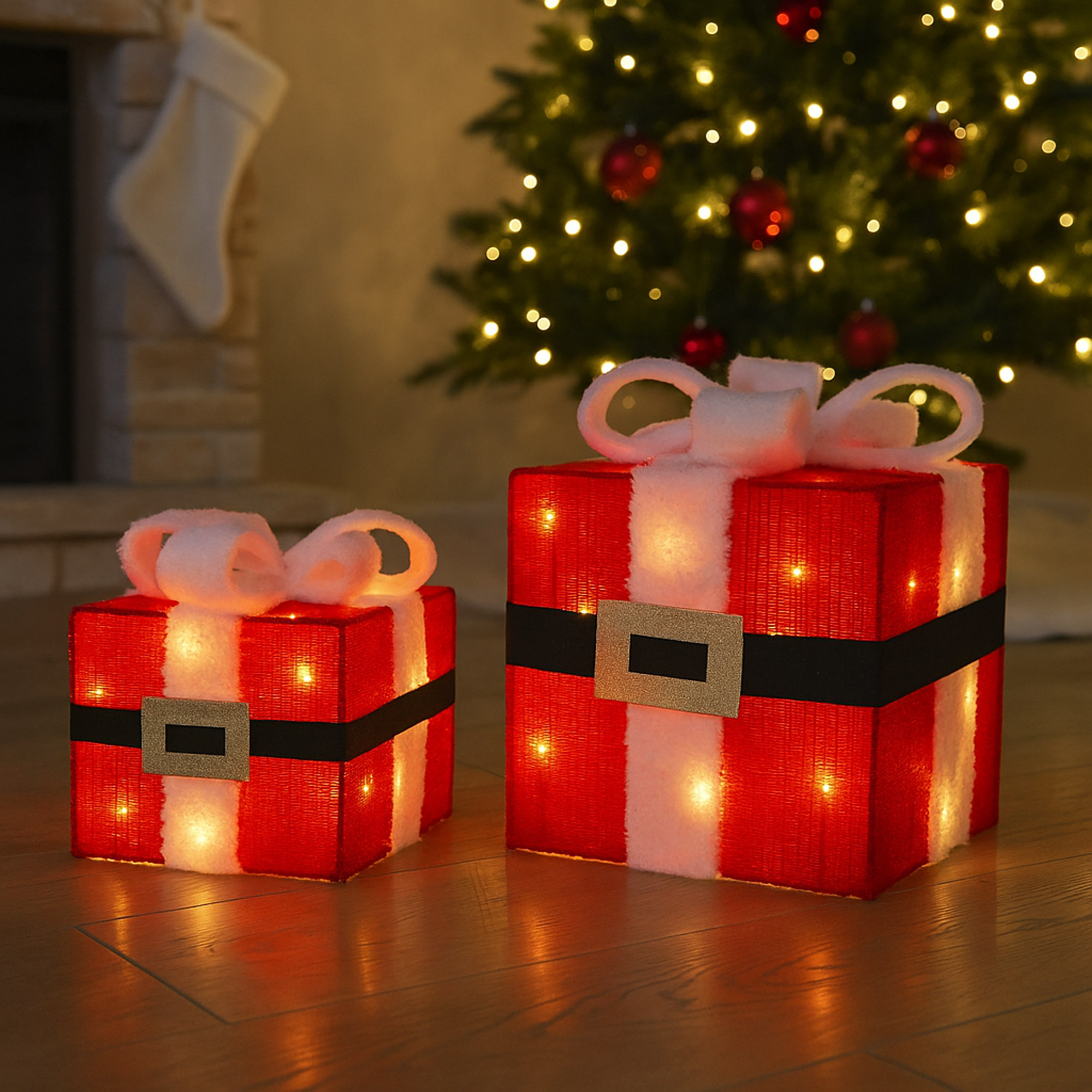 Dos cajas de regalo navideñas iluminadas. Rojas con ribete blanco, cinturón negro, hebilla dorada y lazo blanco. Árbol de Navidad al fondo.