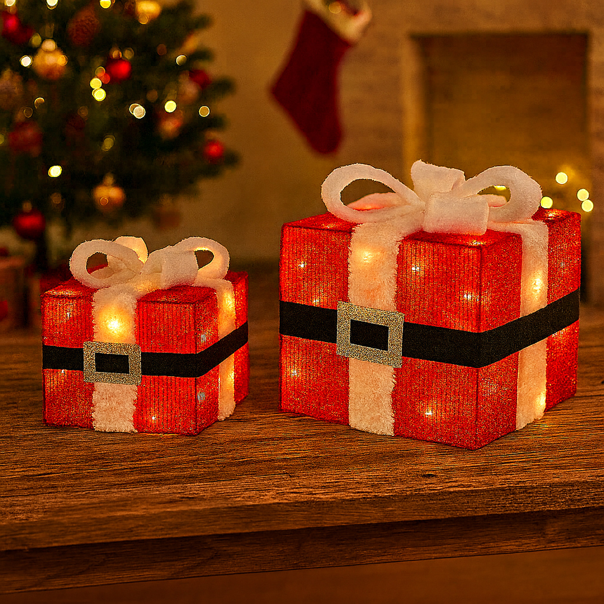 Dos cajas de regalo navideñas iluminadas. Rojas con borde blanco, cinturón negro y hebilla dorada. Un árbol de Navidad borroso al fondo.