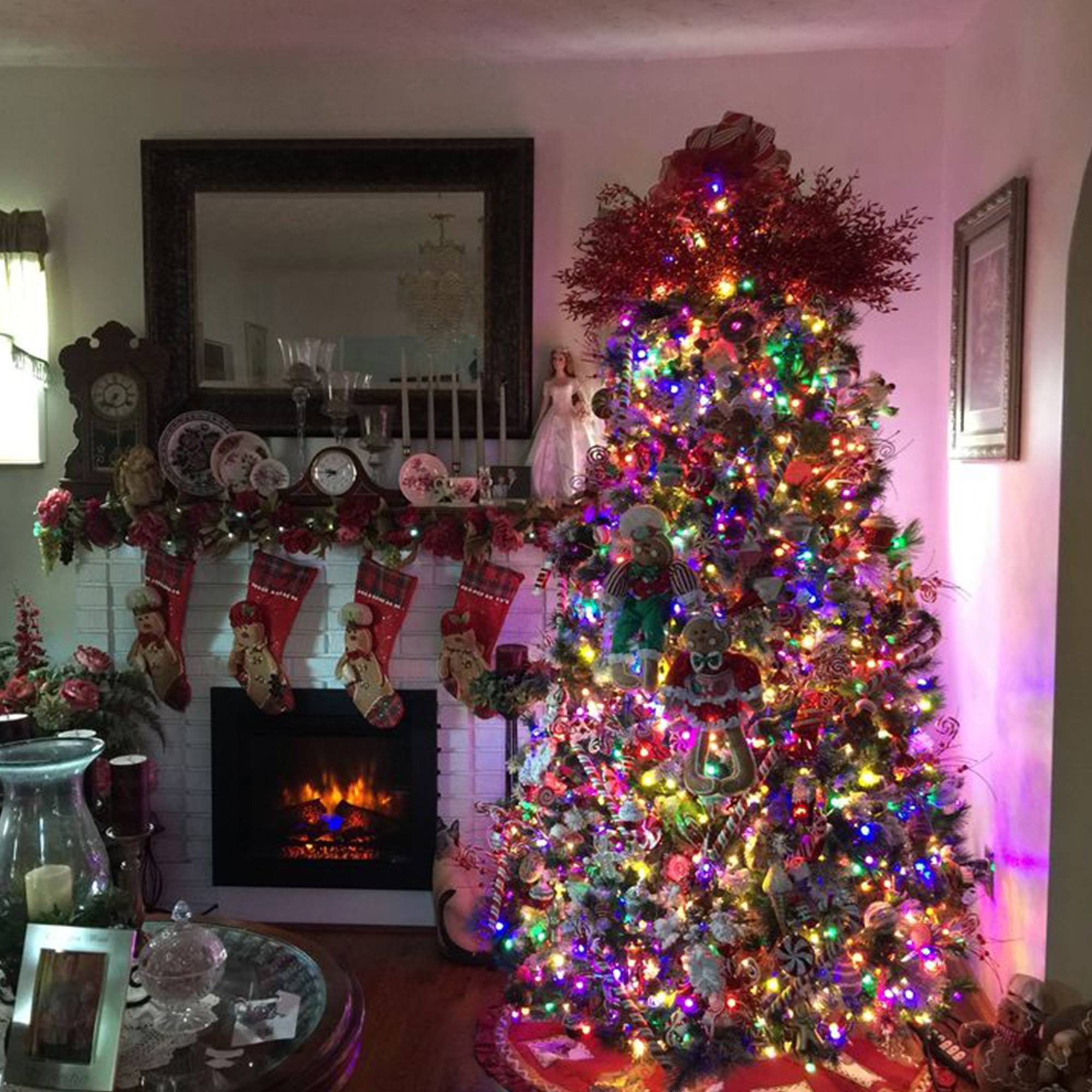 Un árbol de Navidad decorado con luces y adornos. Una chimenea con calcetines, un espejo y una mesa con decoraciones.