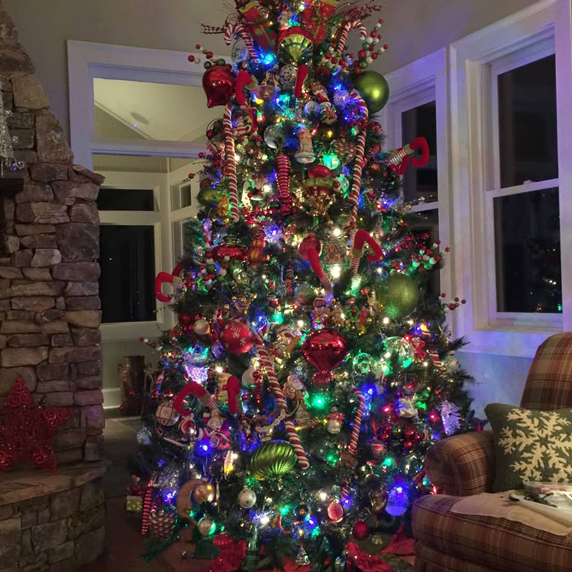 Un árbol de Navidad decorado con adornos y luces, junto a una chimenea de piedra y un sillón a cuadros.