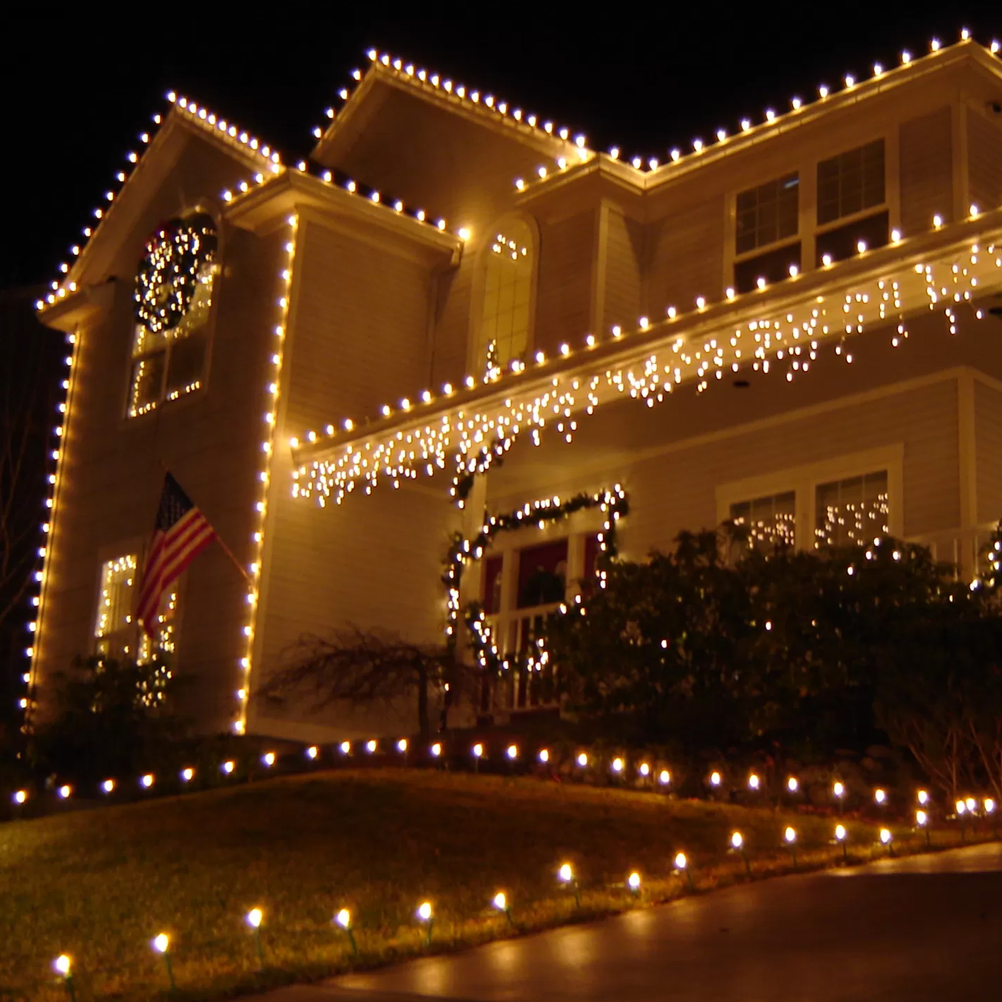 Una casa por la noche, decorada con luces. Una bandera americana cuelga de una ventana. El tejado y los arbustos también están iluminados.